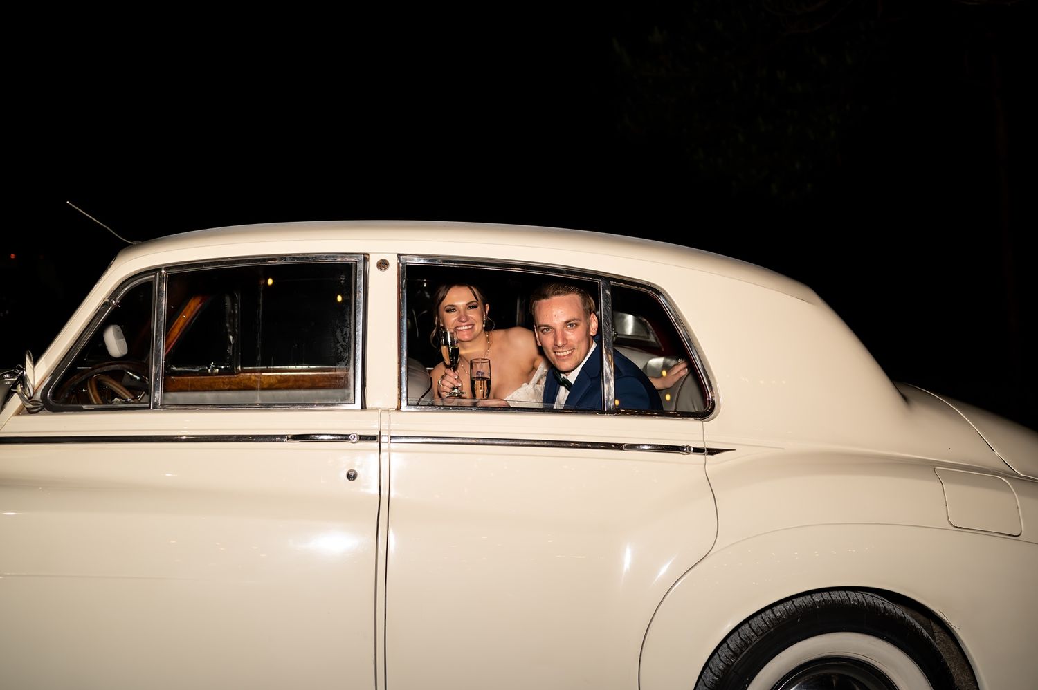 A classic cream-colored vintage car illuminated at night with figures visible through the rear window.