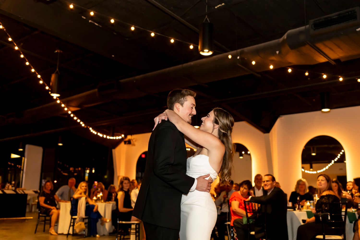 Wedding guests dance beneath warm string lights in an intimate indoor reception venue during evening celebrations.