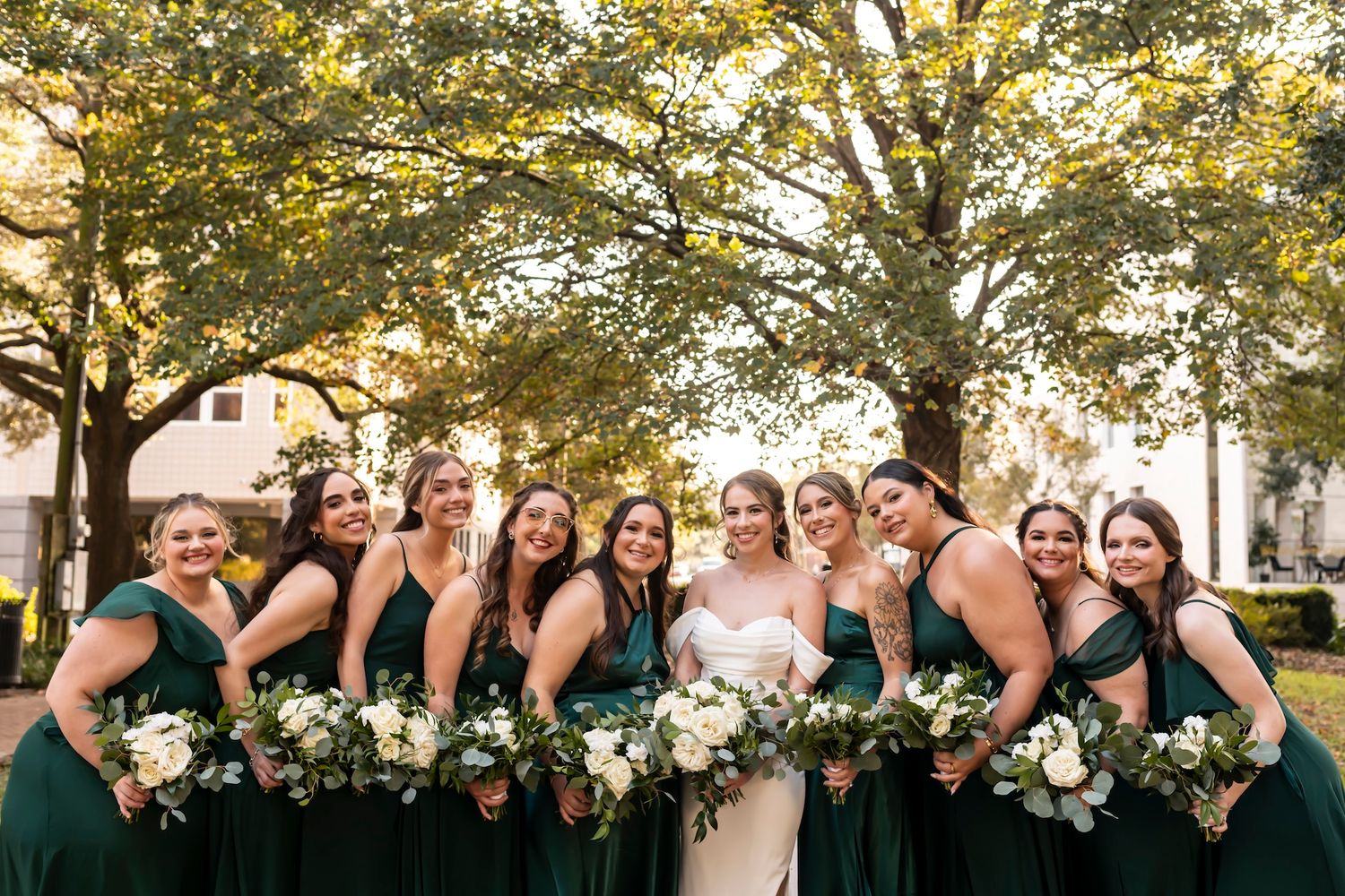 Wedding party wearing emerald green dresses poses together outdoors with white and green bouquets under autumn trees.