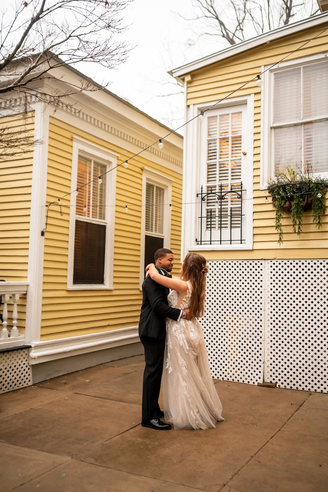 A couple in wedding attire embraces outside a charming yellow historic home with white trim and lattice details.
