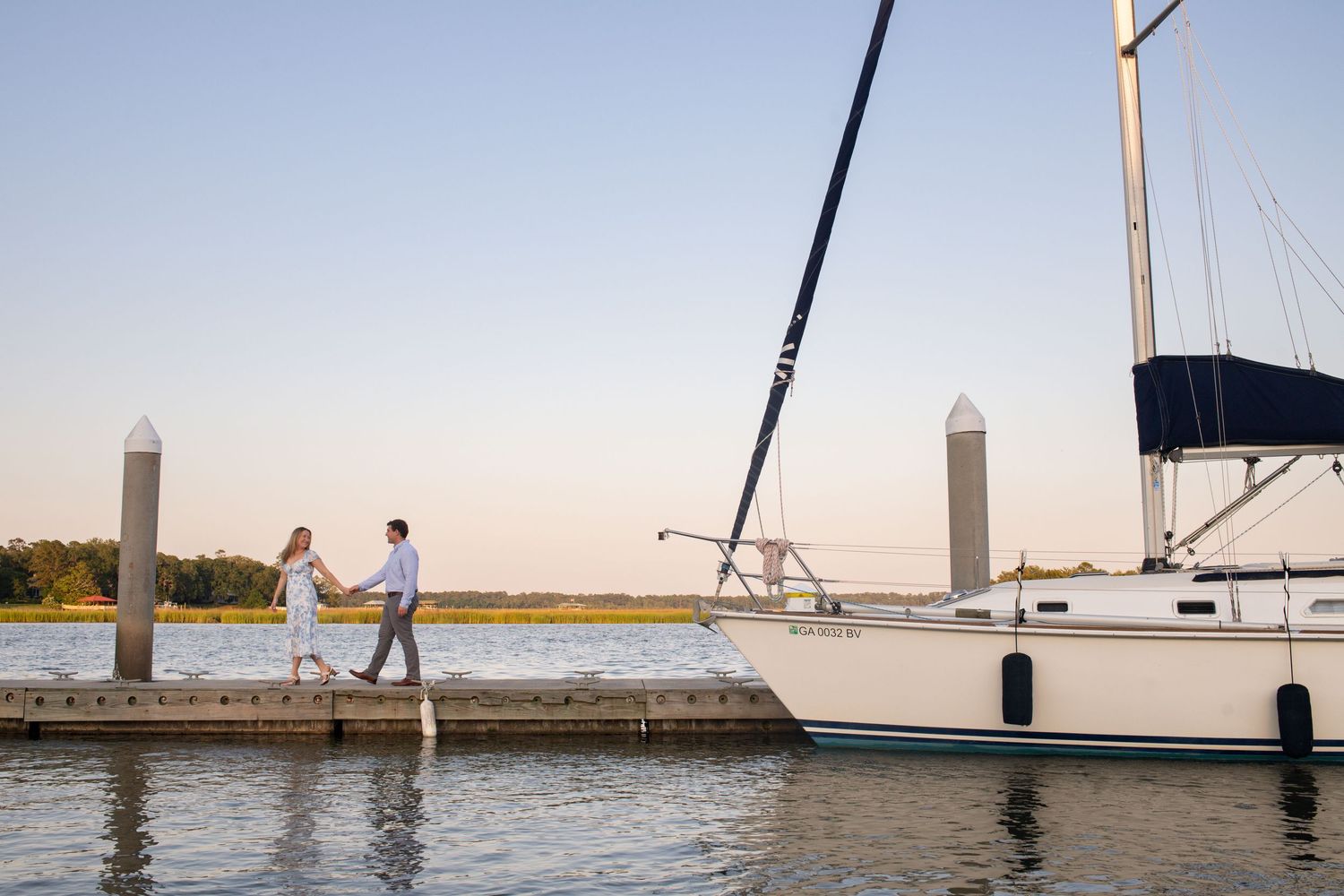 A couple walks along a wooden dock at sunset with a sailboat moored beside concrete pilings in the marina.