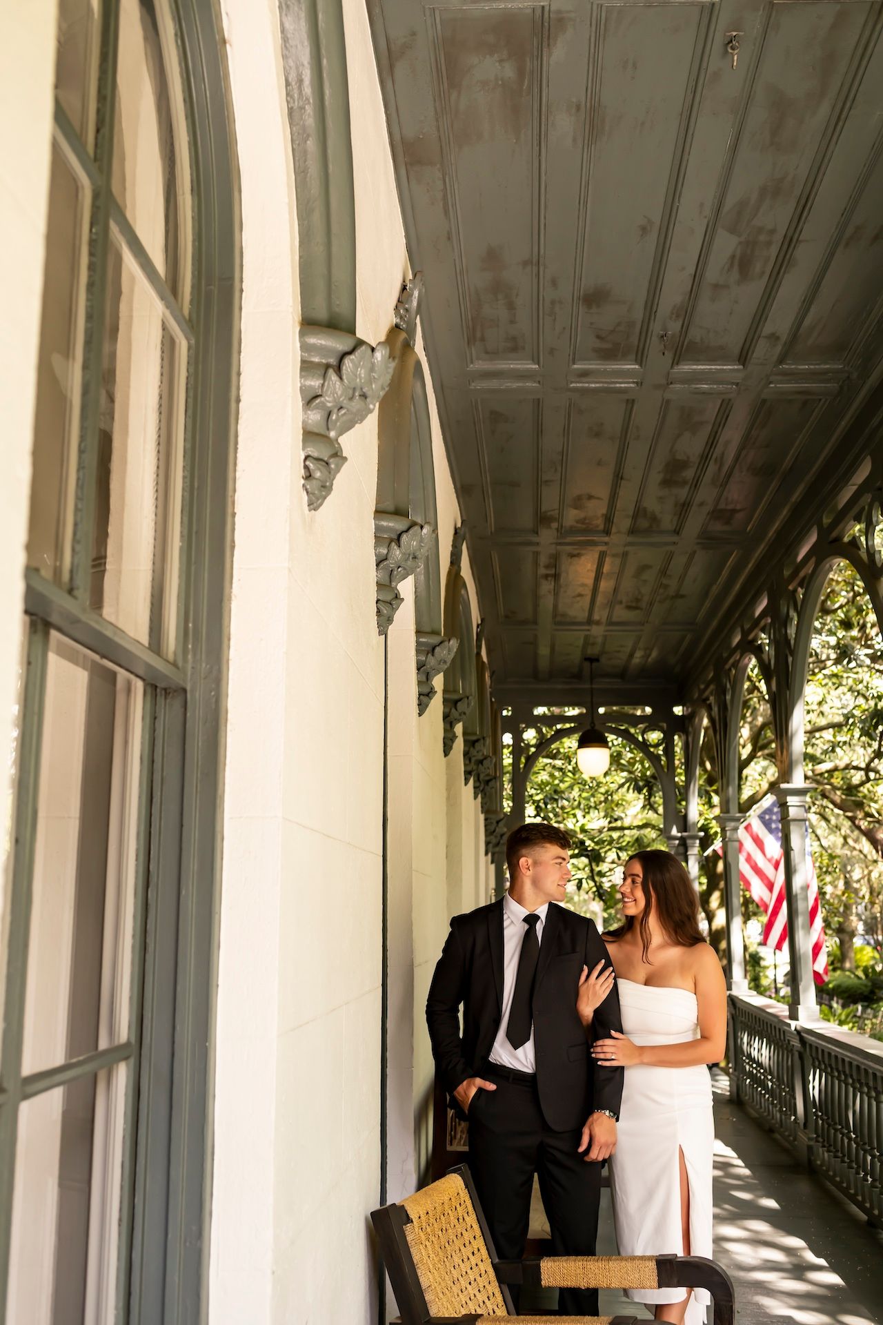 Wedding couple shares a romantic moment in a white-pillared columned porch walkway with dappled sunlight.