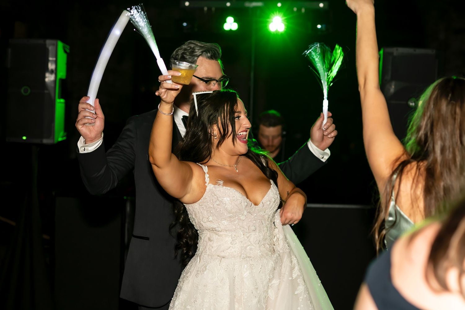 Party guests hold glow sticks in the air during a lively wedding reception celebration under green lighting.