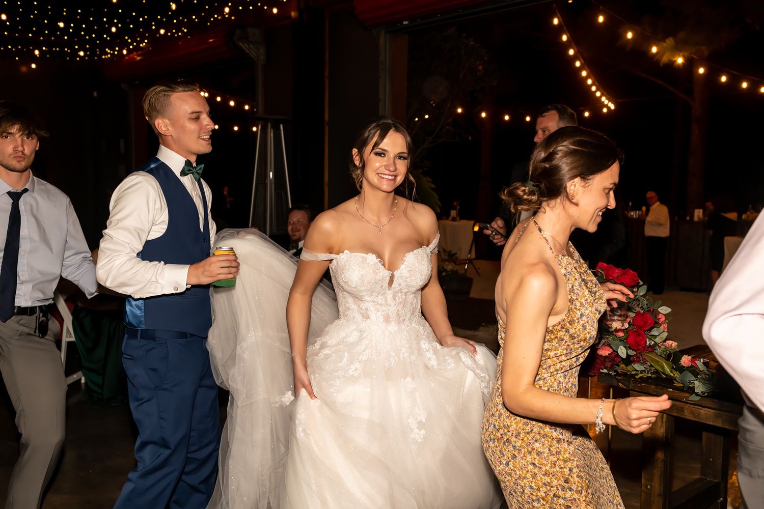 Wedding party members dancing and celebrating at an evening reception under string lights.