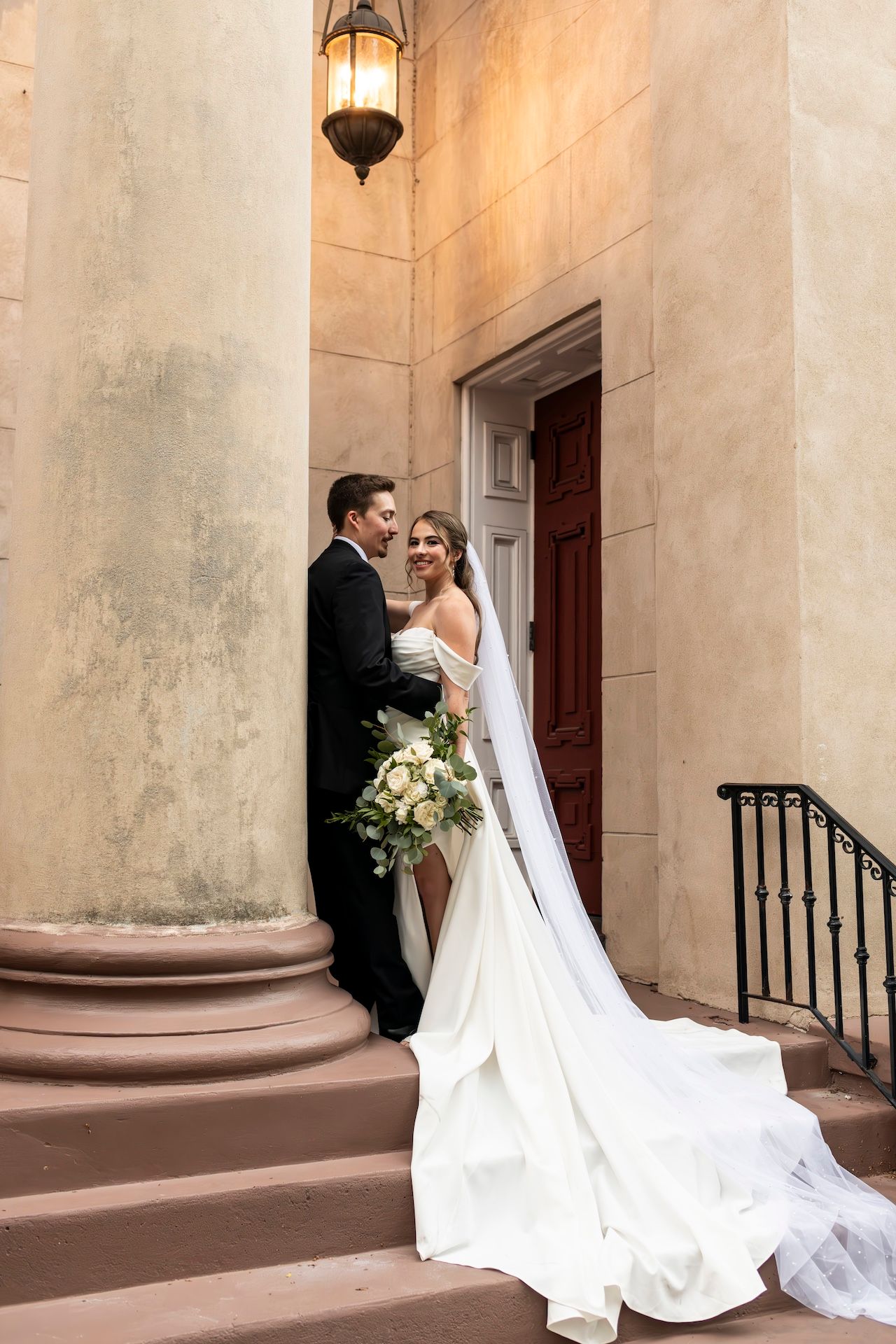 A couple embraces on stone steps by a columned entrance with an elegant white wedding gown trailing behind.
