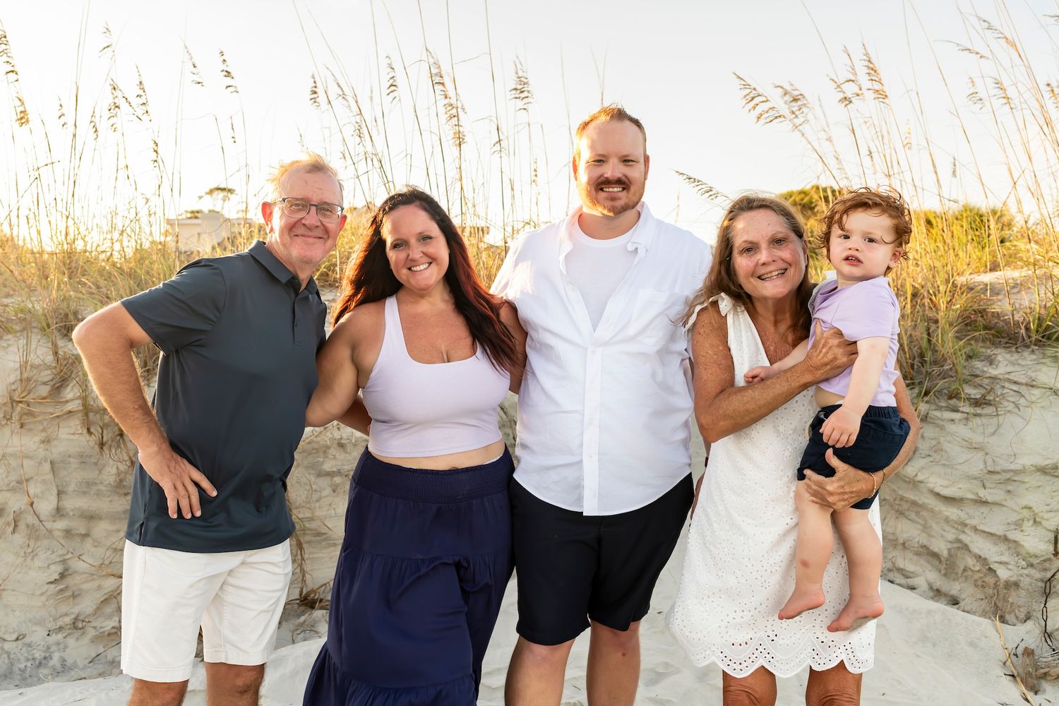 Group poses together on a sunny beach with sand dunes and tall grass in the background at golden hour.