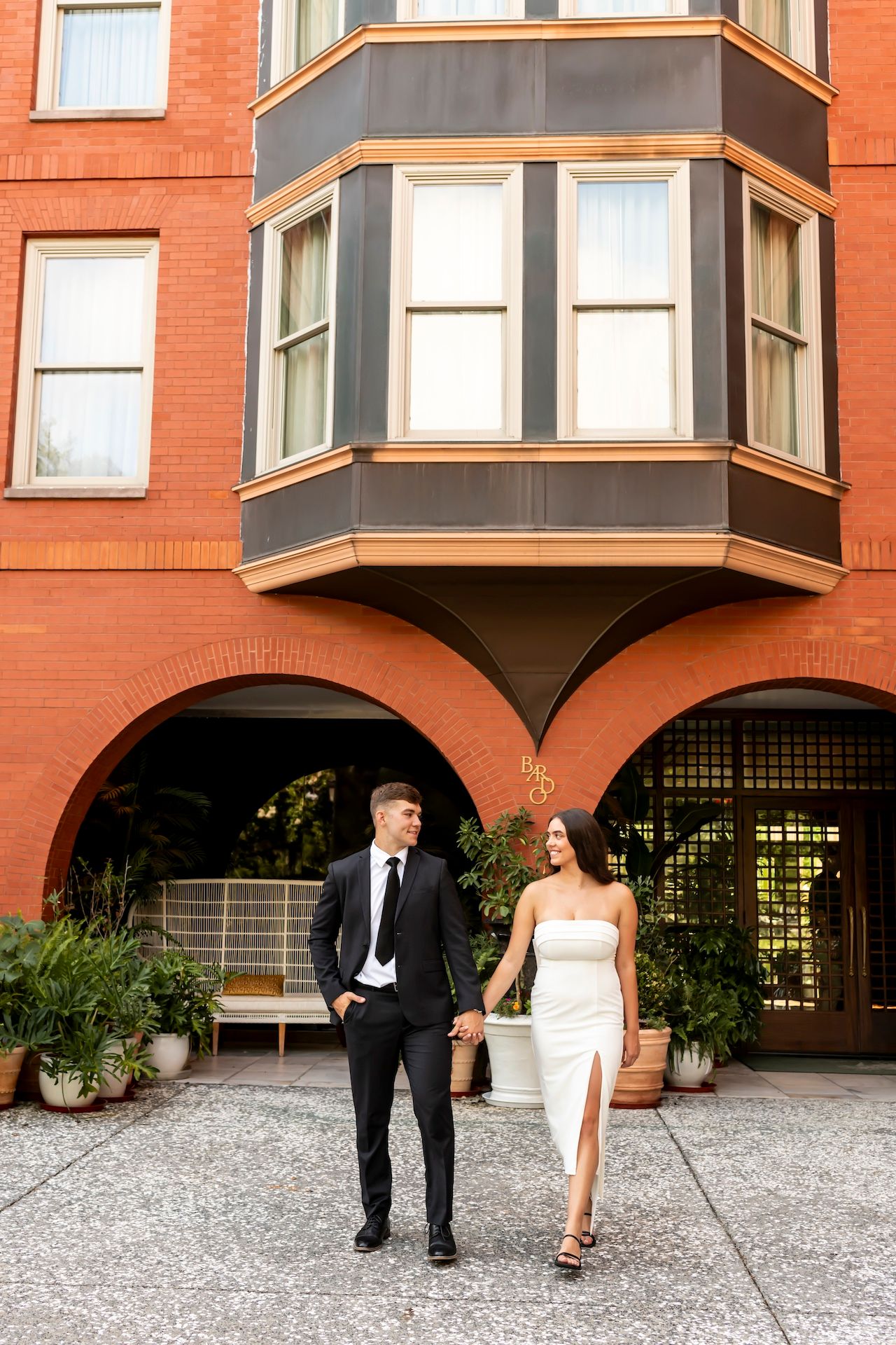 A couple in formal attire walking together outside a red brick building with a distinctive bay window.