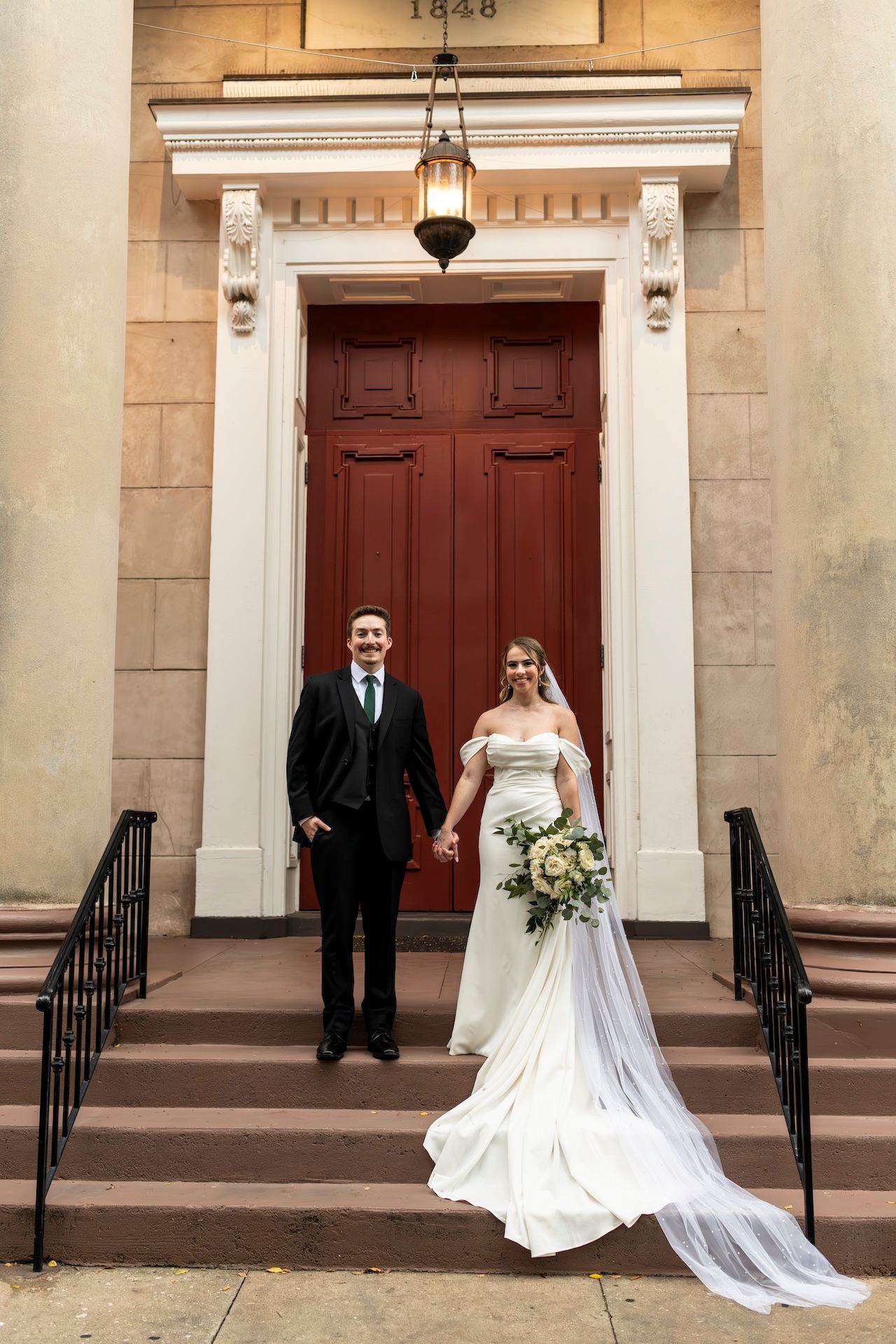 Wedding couple poses in front of stone church building with red wooden doors and white columns in elegant ceremony photos.