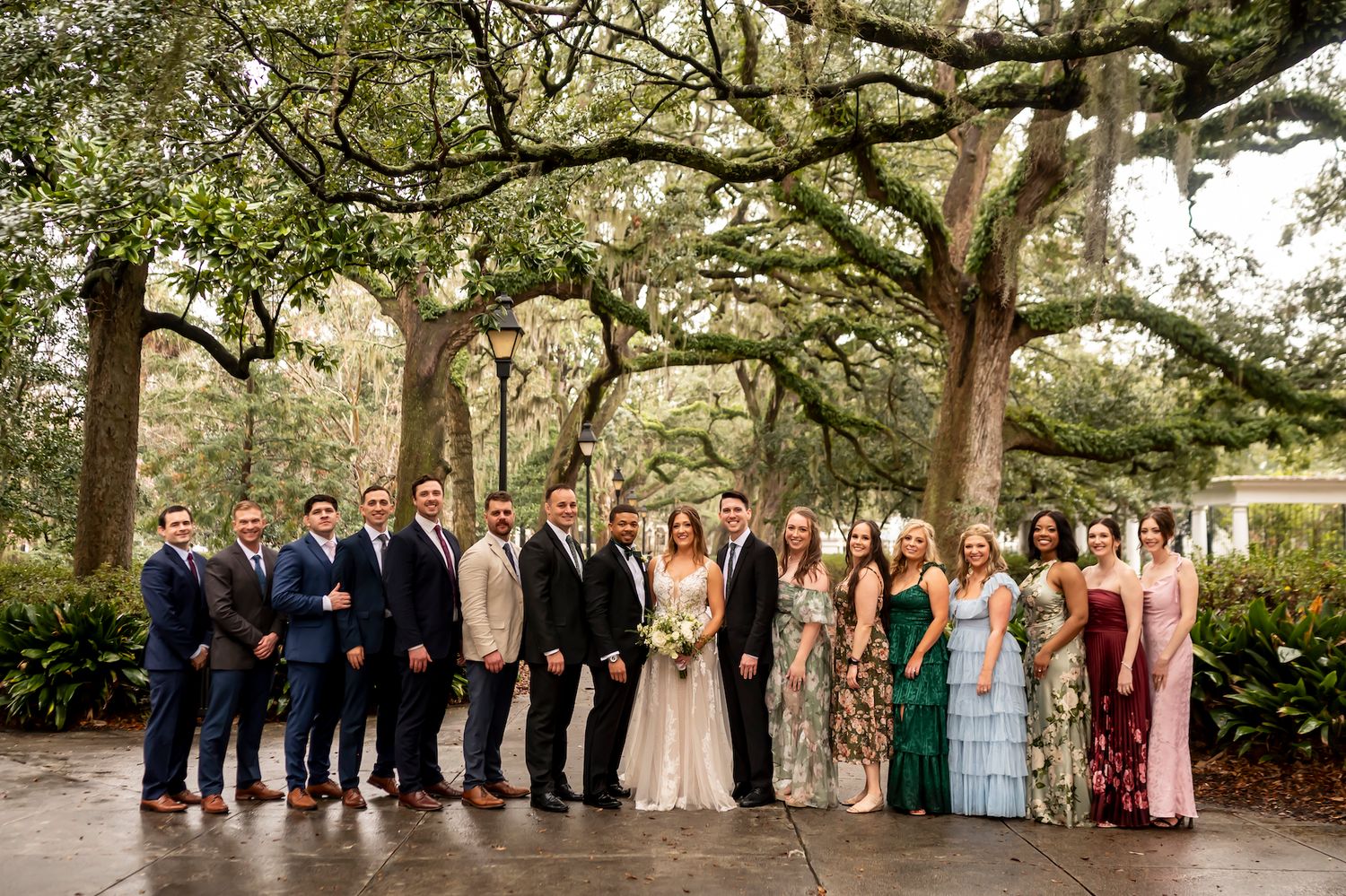 A wedding party poses under majestic oak trees with Spanish moss on a cloudy day in a garden setting.