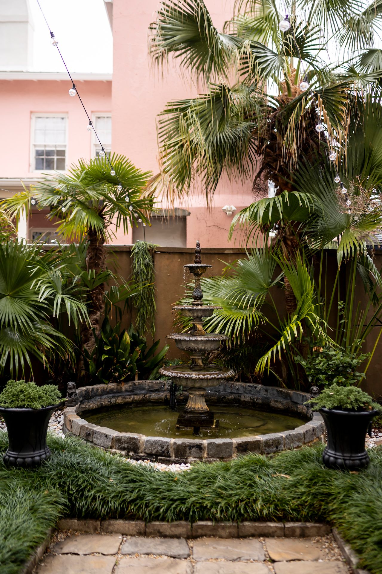 A tropical garden courtyard with palm trees and stone fountain surrounded by lush greenery against a coral pink building.