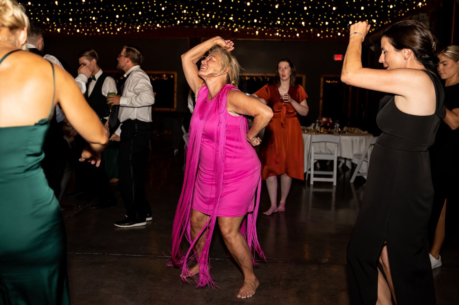 Dancers in bright pink and green dresses move energetically on a dimly lit dance floor during an evening celebration.