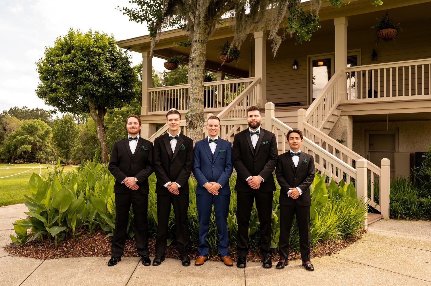 Five groomsmen in formal attire stand in front of steps at an outdoor venue with lush landscaping.