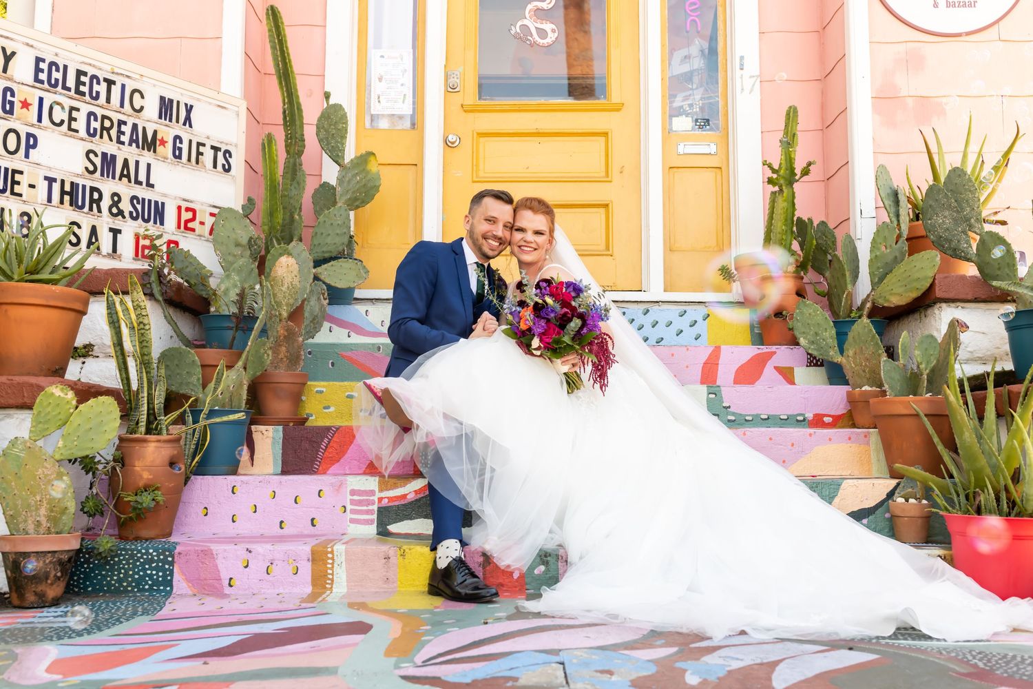 Colorful party decorations including balloons and paper ornaments adorn a pink and yellow space with potted cactus plants.