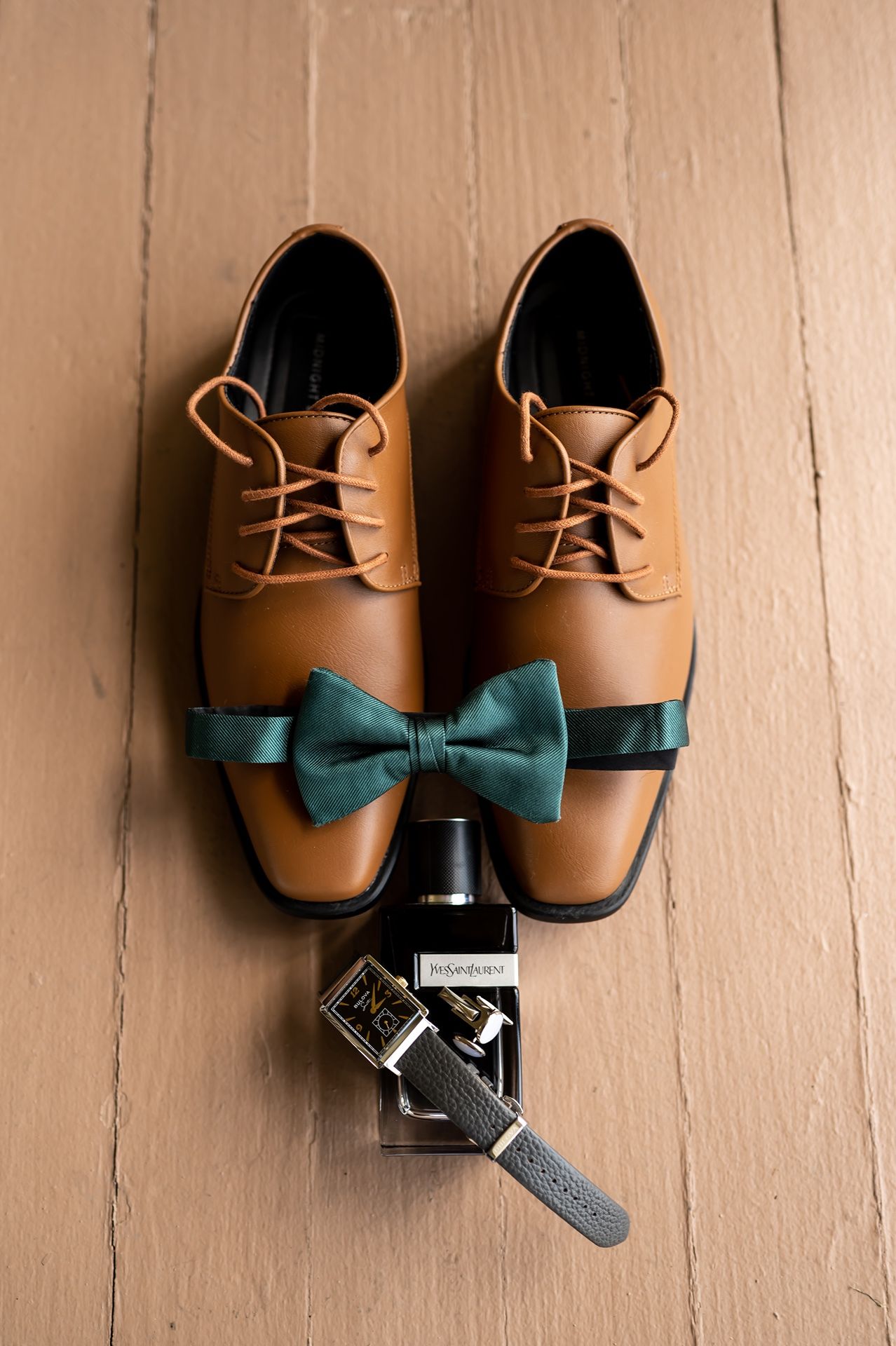 Brown leather dress shoes with teal bow tie and vintage cologne bottle arranged on wooden floor.