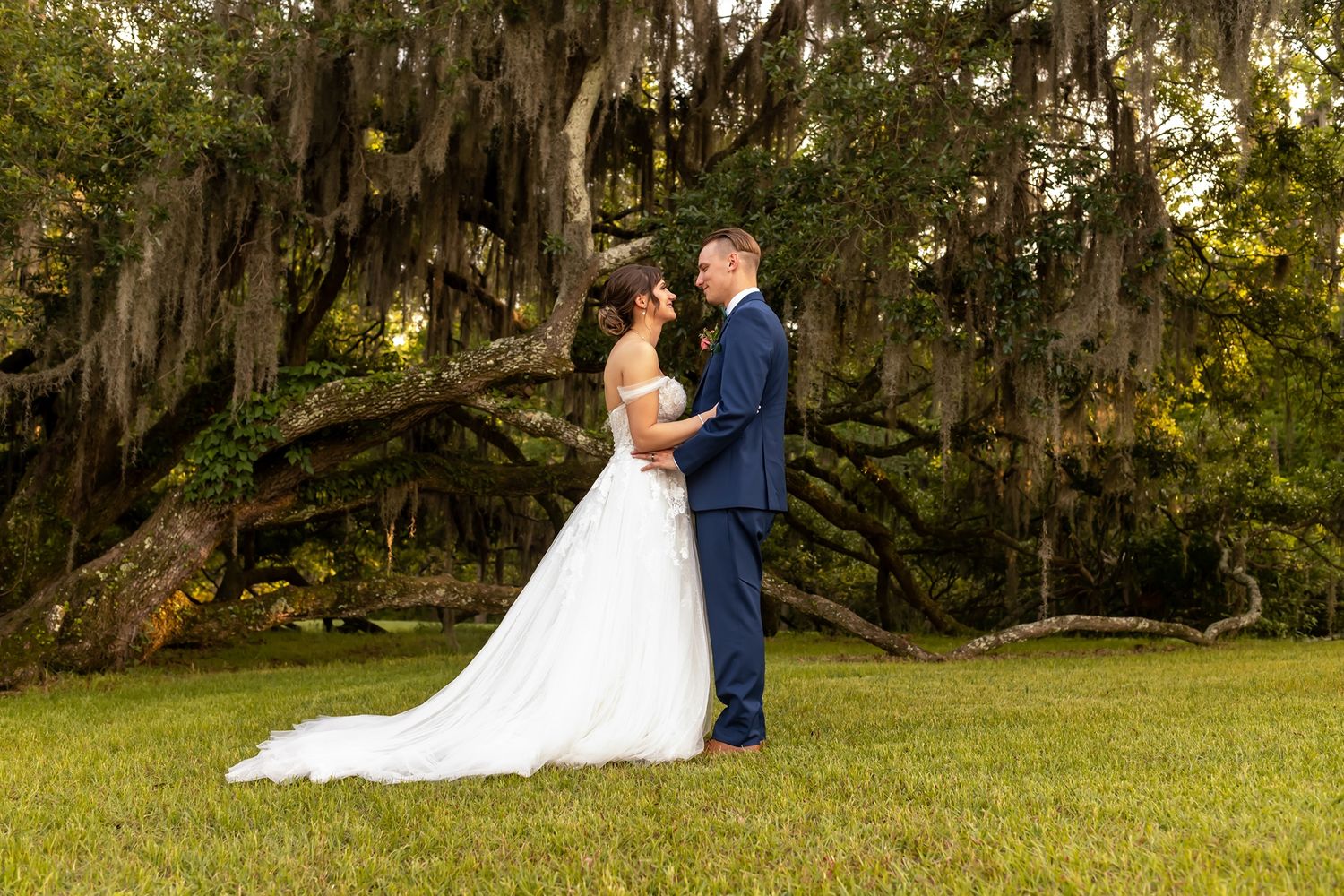 Two figures embrace during an outdoor wedding ceremony under moss-draped oak trees on a sunny day.