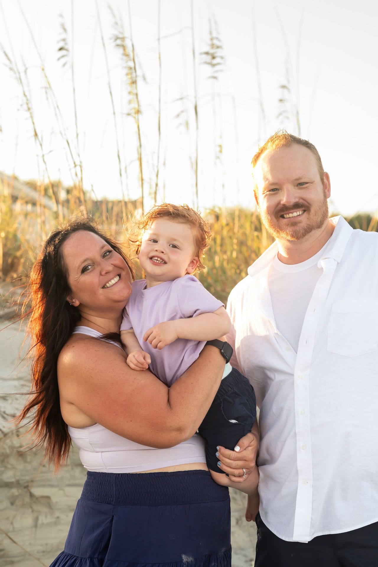 A family shares a joyful moment together on a sandy beach during golden hour with tall grass in the background.