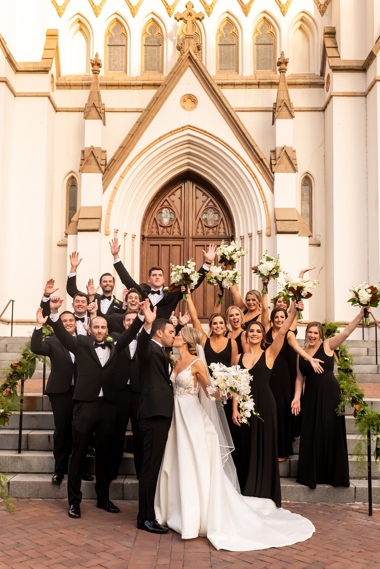 Wedding party in formal black and white attire celebrates joyfully outside ornate Gothic church architecture.