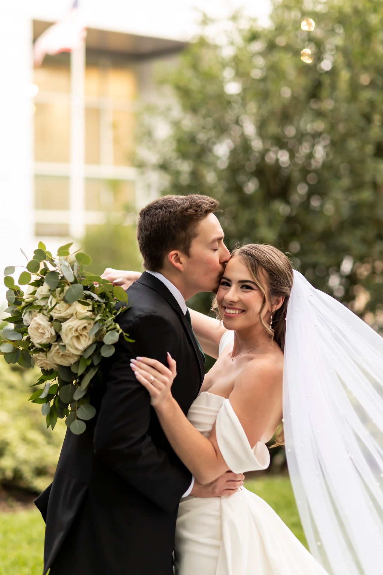 Romantic wedding moment captured as couple embraces outdoors with white roses and greenery bouquet.