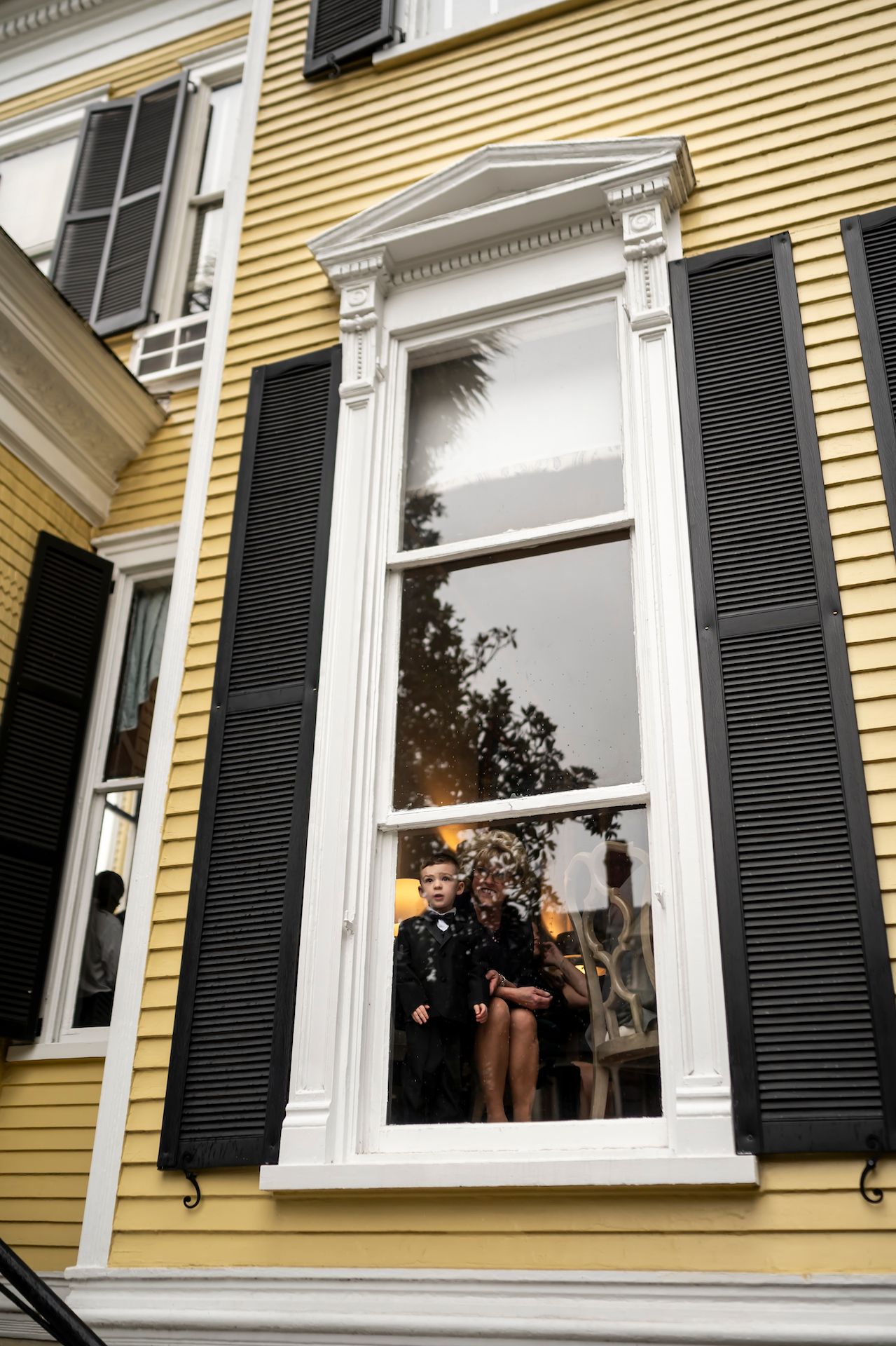 Tall window with ornate white trim and black shutters on a yellow historic house shows reflections at sunset.