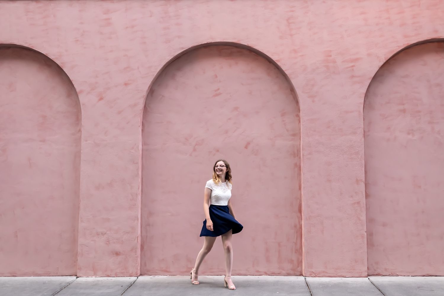 A person in a flowy dress poses elegantly against a pink wall with arched architectural details.