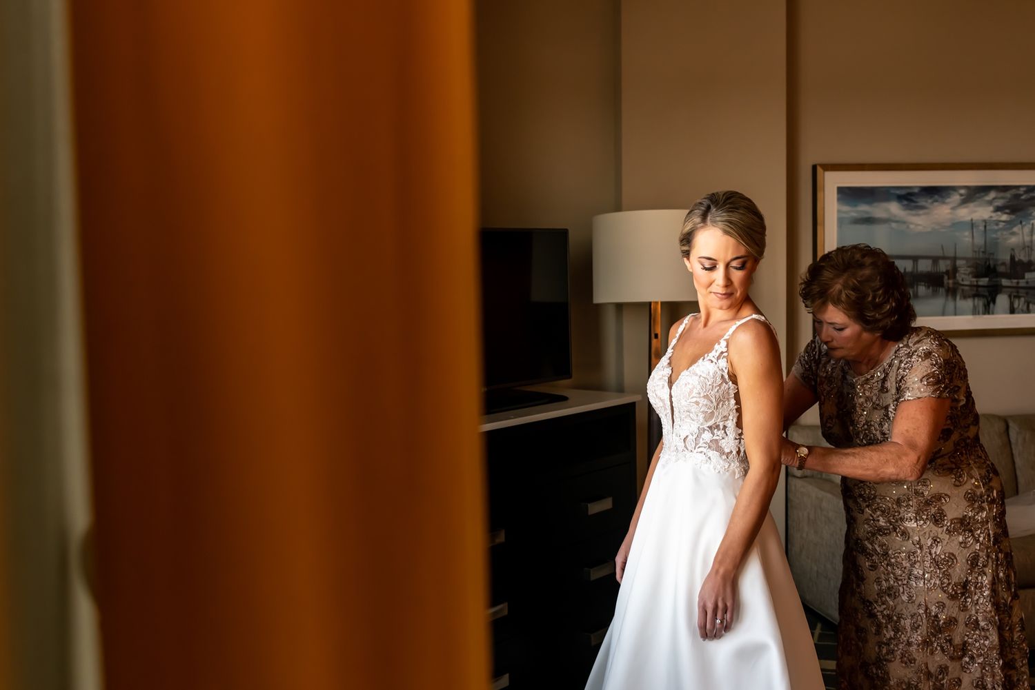 A bride in a white lace gown gets help with final adjustments to her dress while standing in a sunlit room.