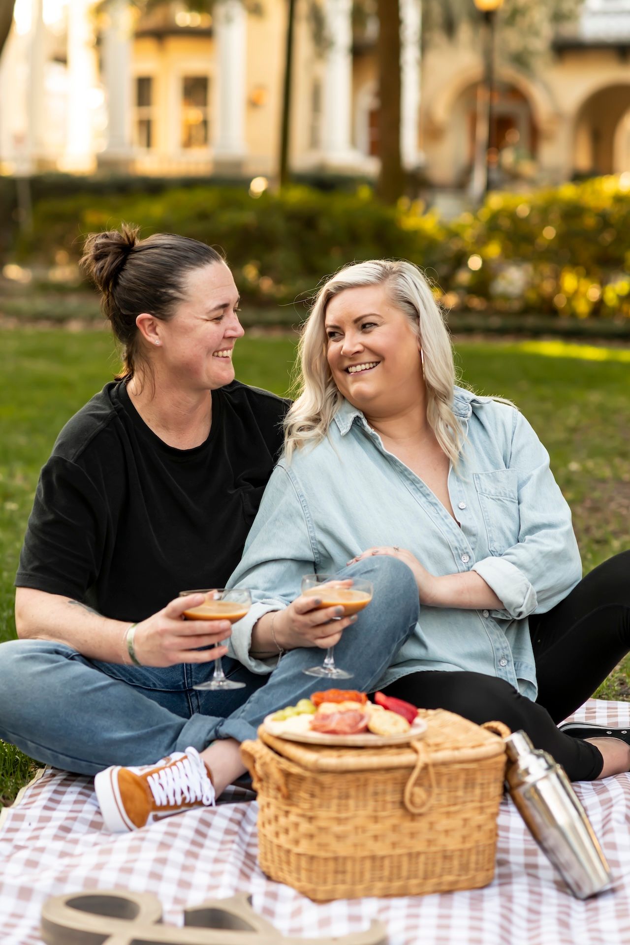 Two friends enjoy wine and snacks during a picnic on a checkered blanket with a wicker basket outdoors.