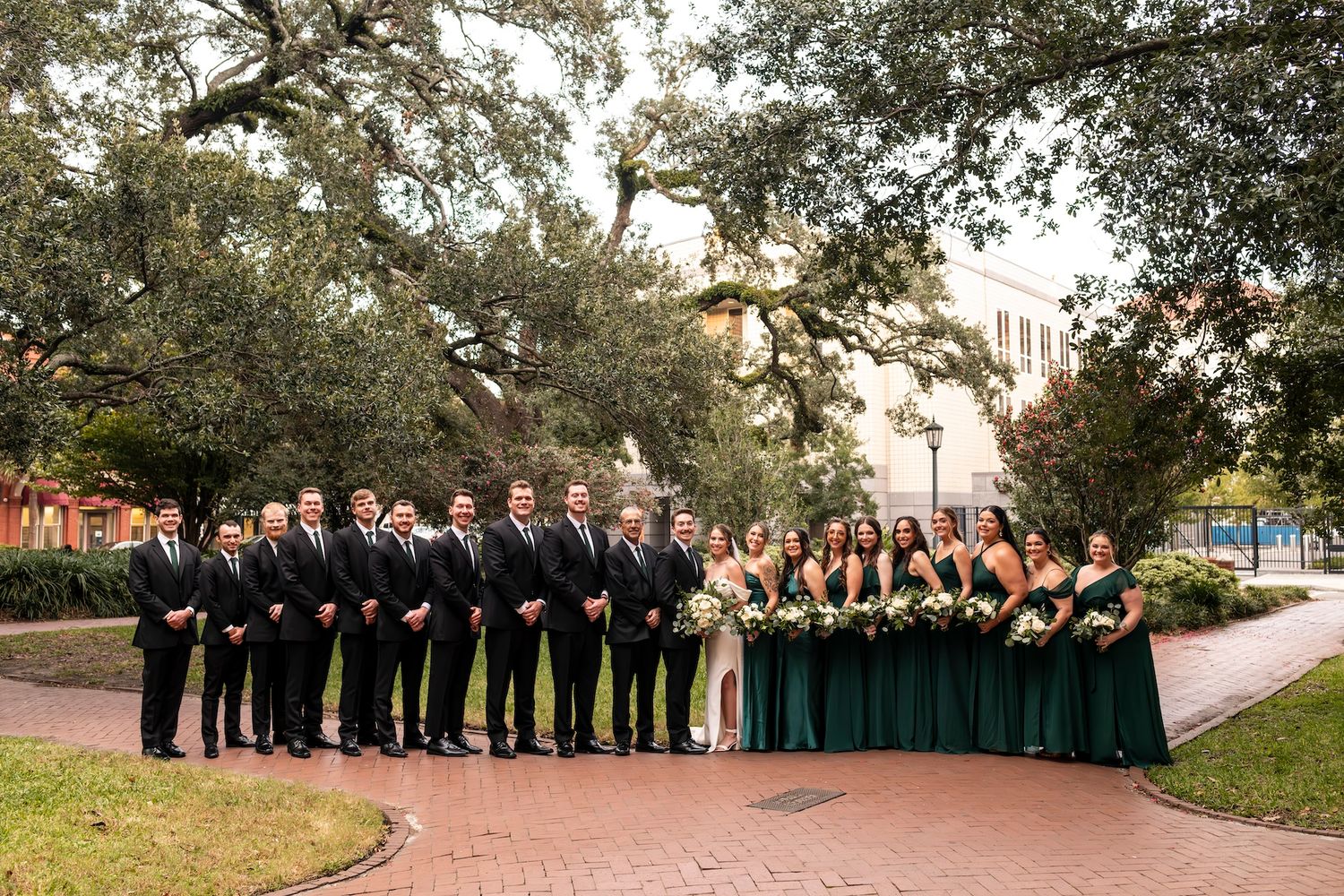 A wedding party in formal attire stands outdoors beneath oak trees on a brick path during golden hour.