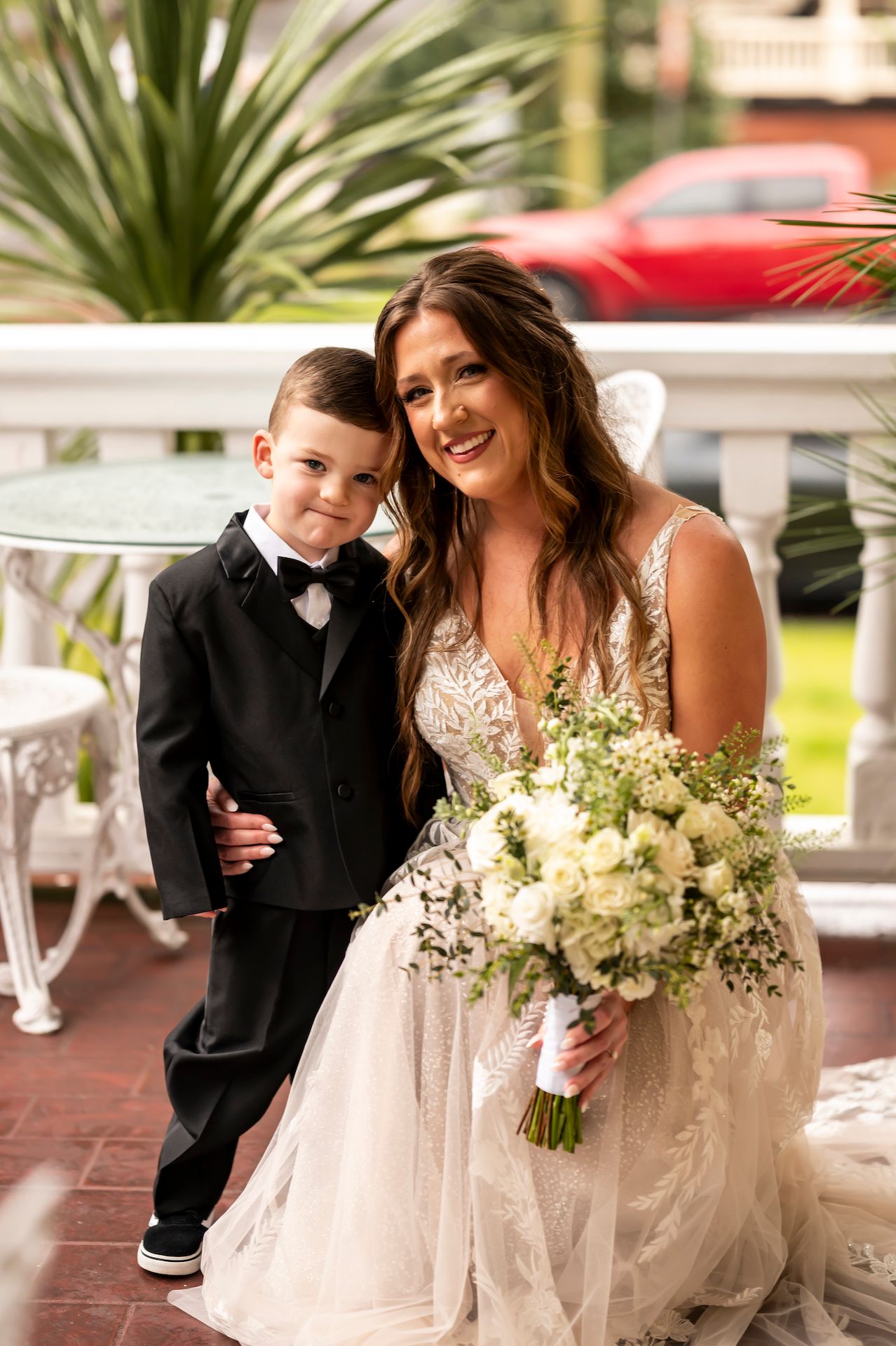 Young ring bearer in black tuxedo poses with bride holding white floral bouquet on outdoor porch.