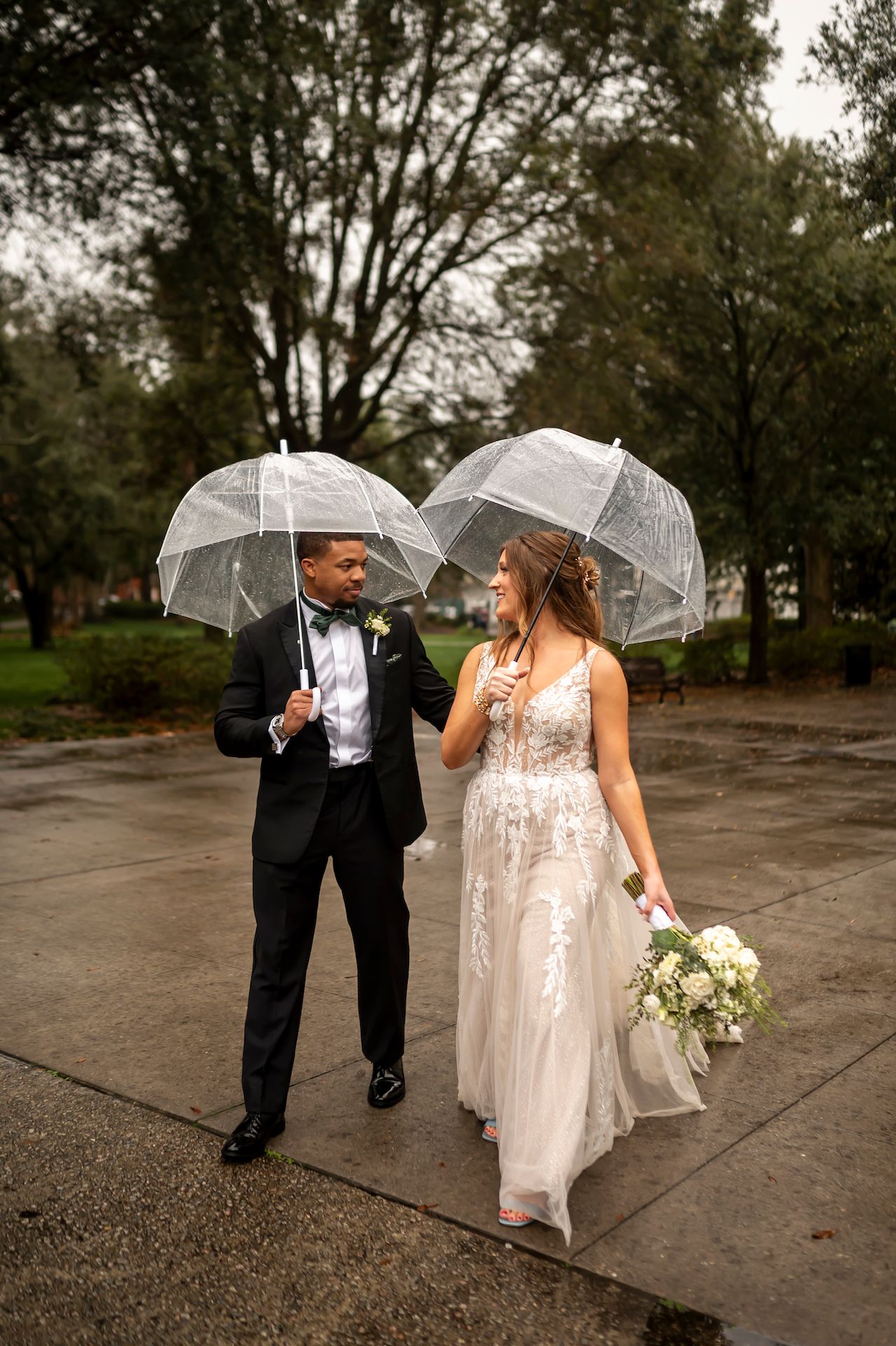 A bride and groom smile at each other while holding clear umbrellas on a rainy day in a park.