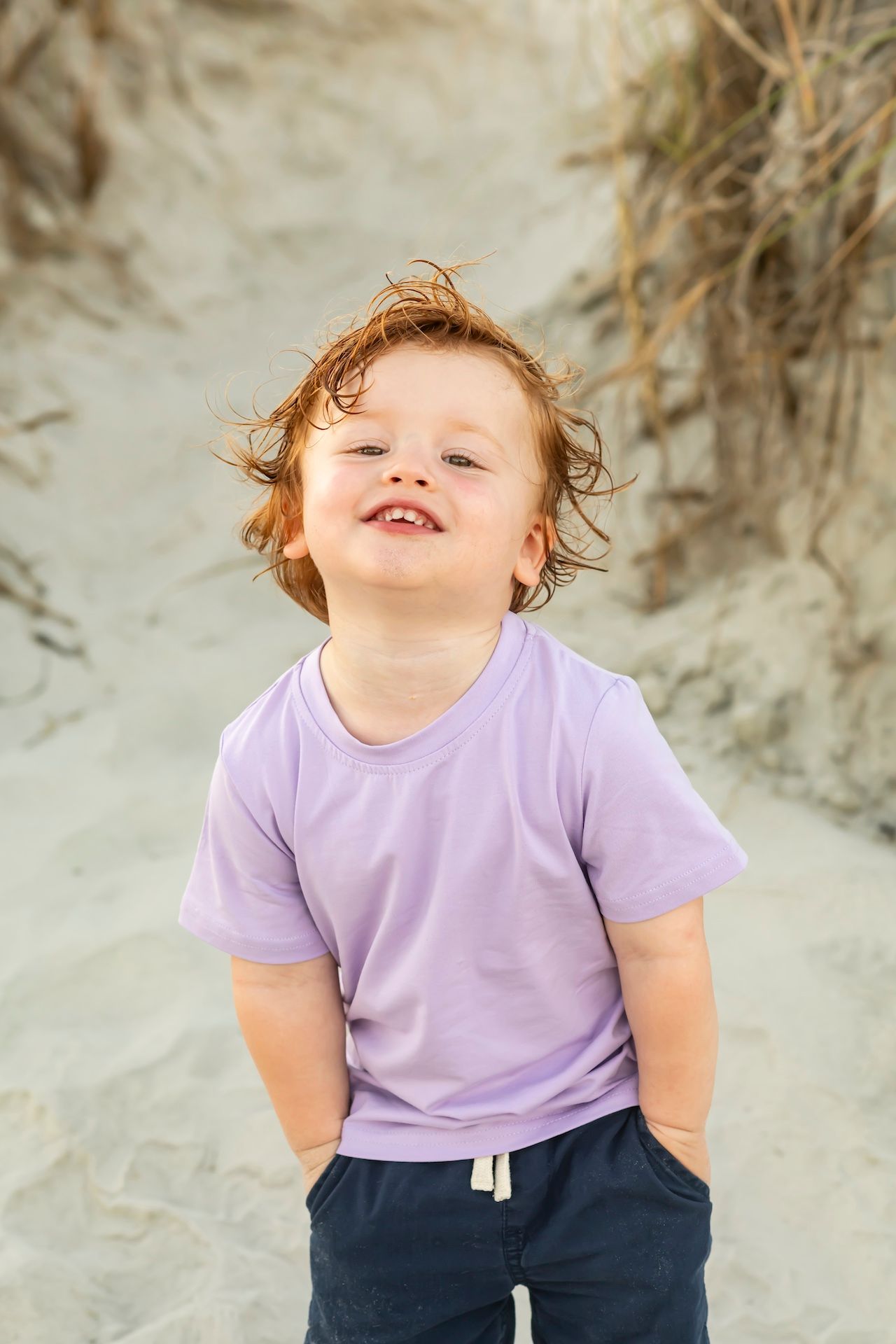 Small child wearing purple t-shirt and navy pants standing on sandy beach with windswept hair.