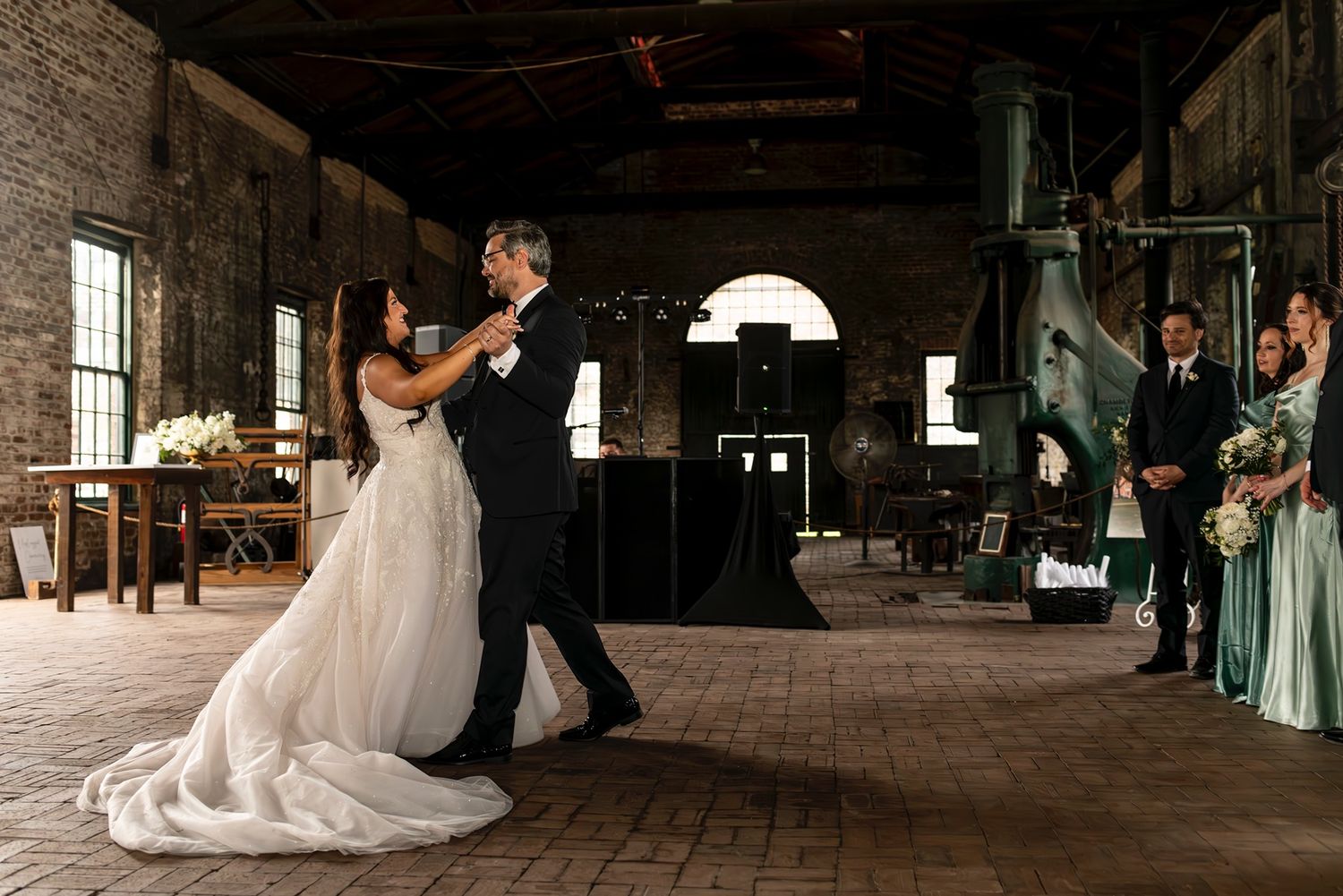 A couple shares their first dance in a rustic industrial venue with brick walls and wooden floors as guests look on.