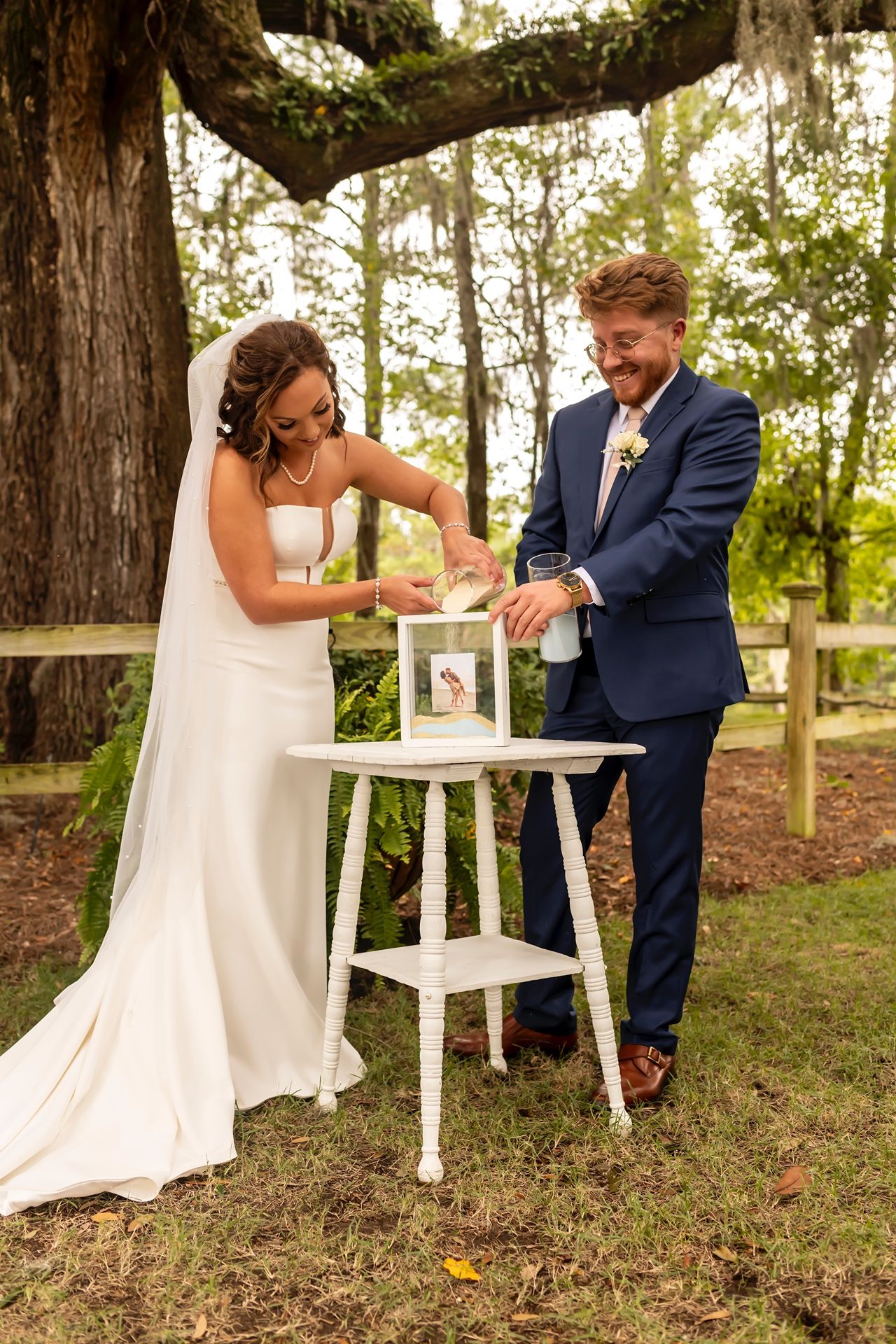 Newlyweds share a joyful moment pouring unity sand into a white decorative stand during their outdoor woodland ceremony.