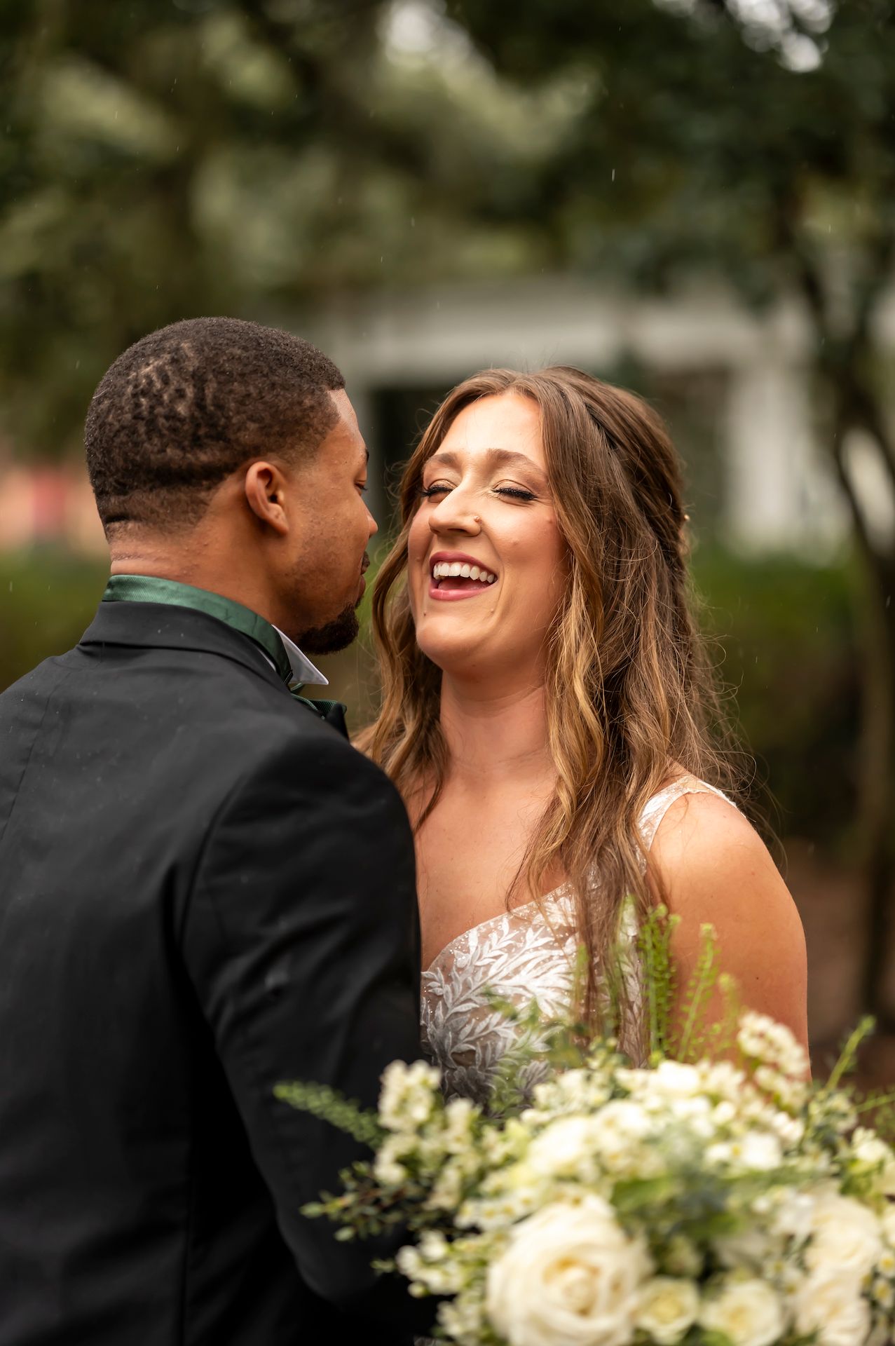 A joyful couple shares an intimate moment amongst greenery while holding a white floral bouquet on their wedding day.