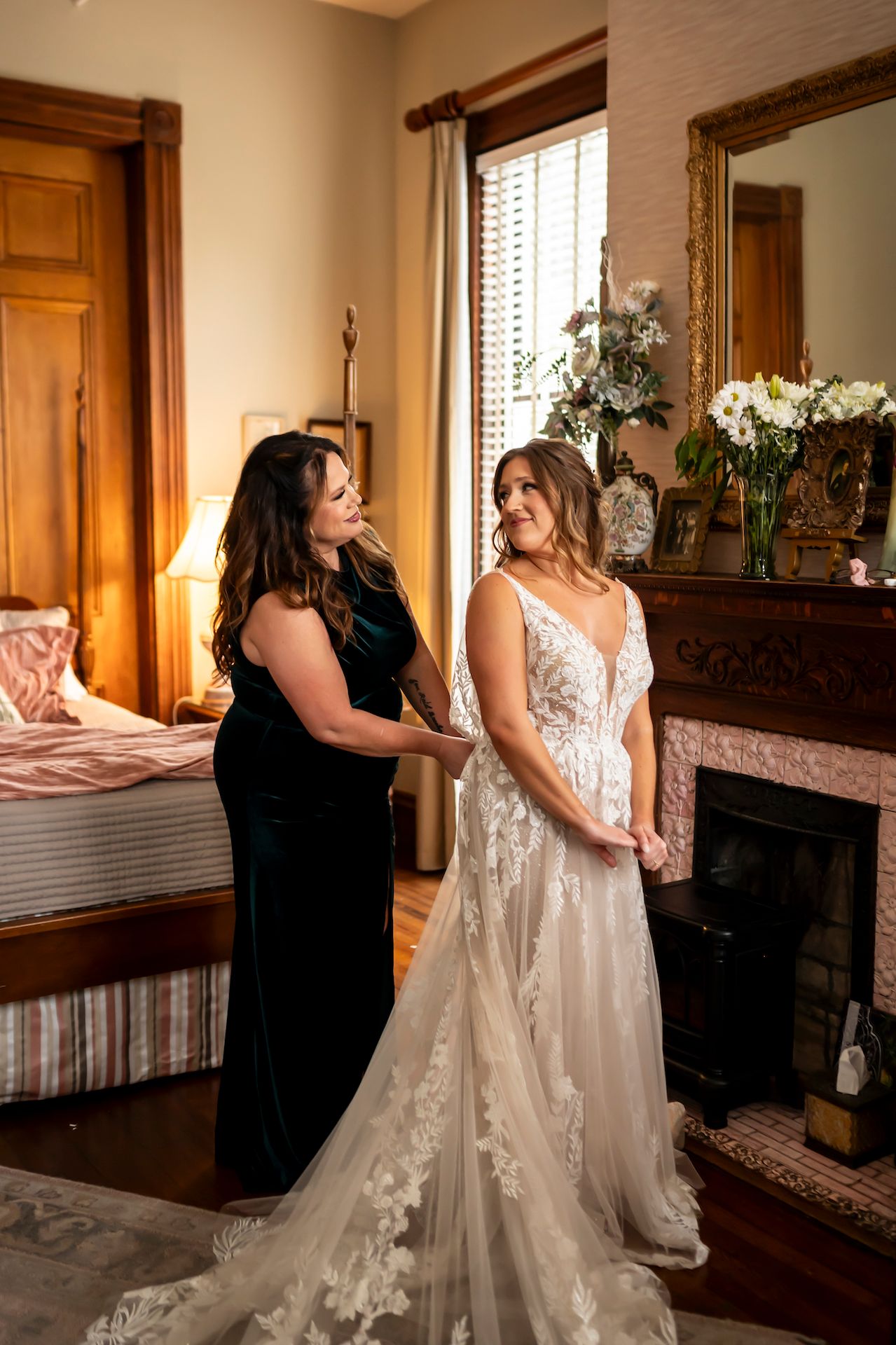Two women in formal dresses stand by a fireplace in an elegant room with one wearing a flowing white wedding gown.