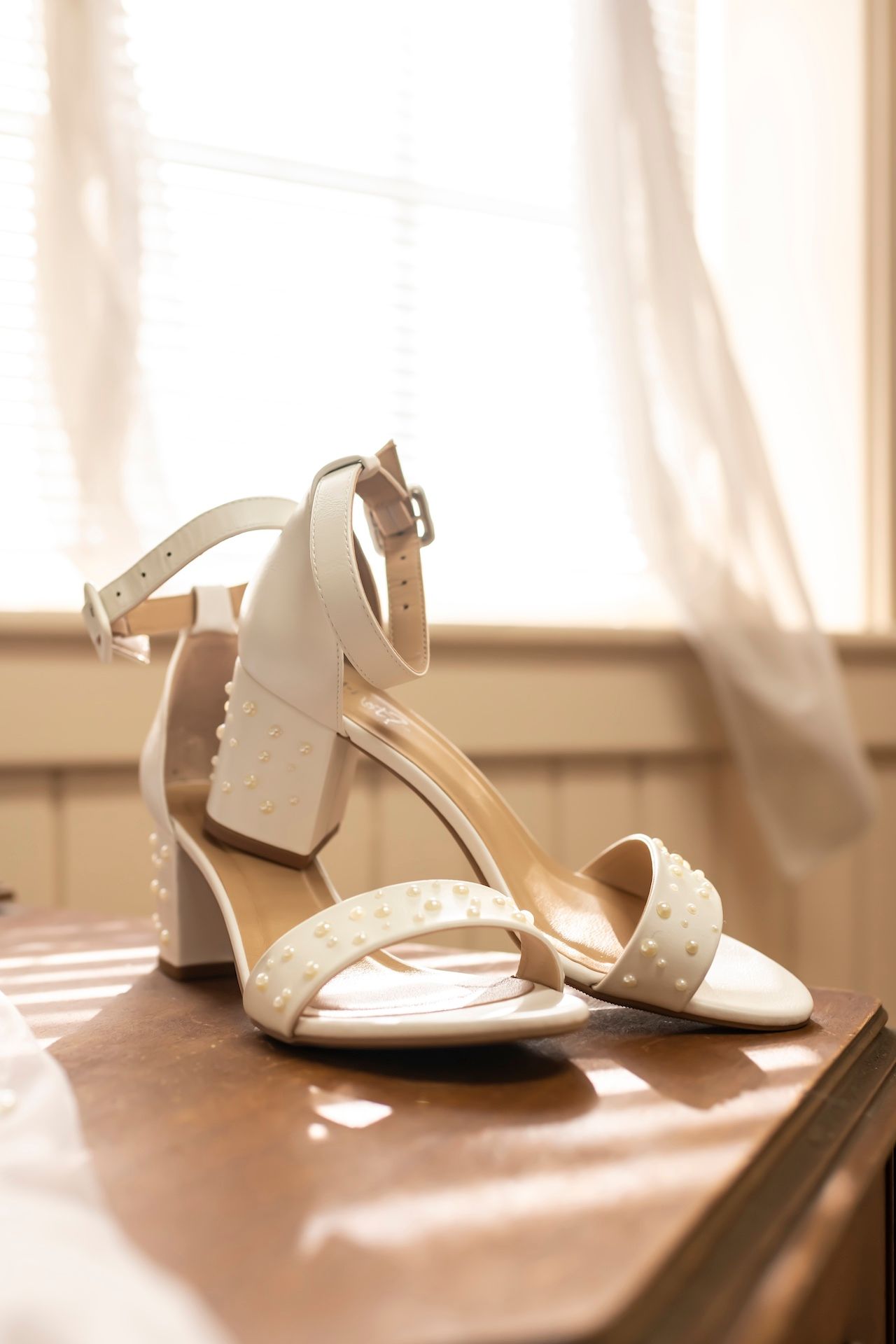 Elegant white wedding sandals with chunky heels and ankle straps sit on a brown surface in soft natural lighting.