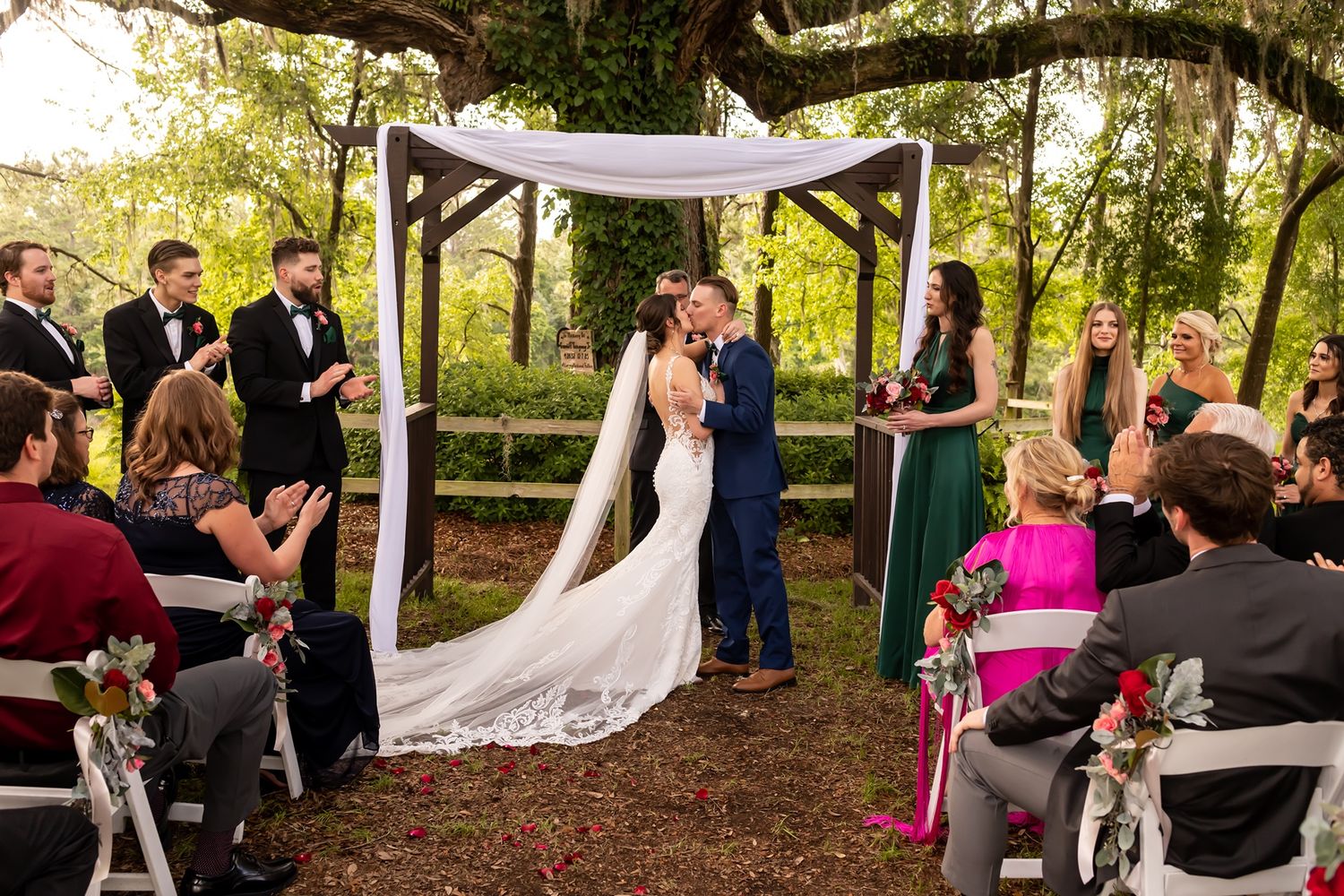Beautiful outdoor wedding ceremony under wooden arbor decorated with white fabric and floral accents in lush garden setting.