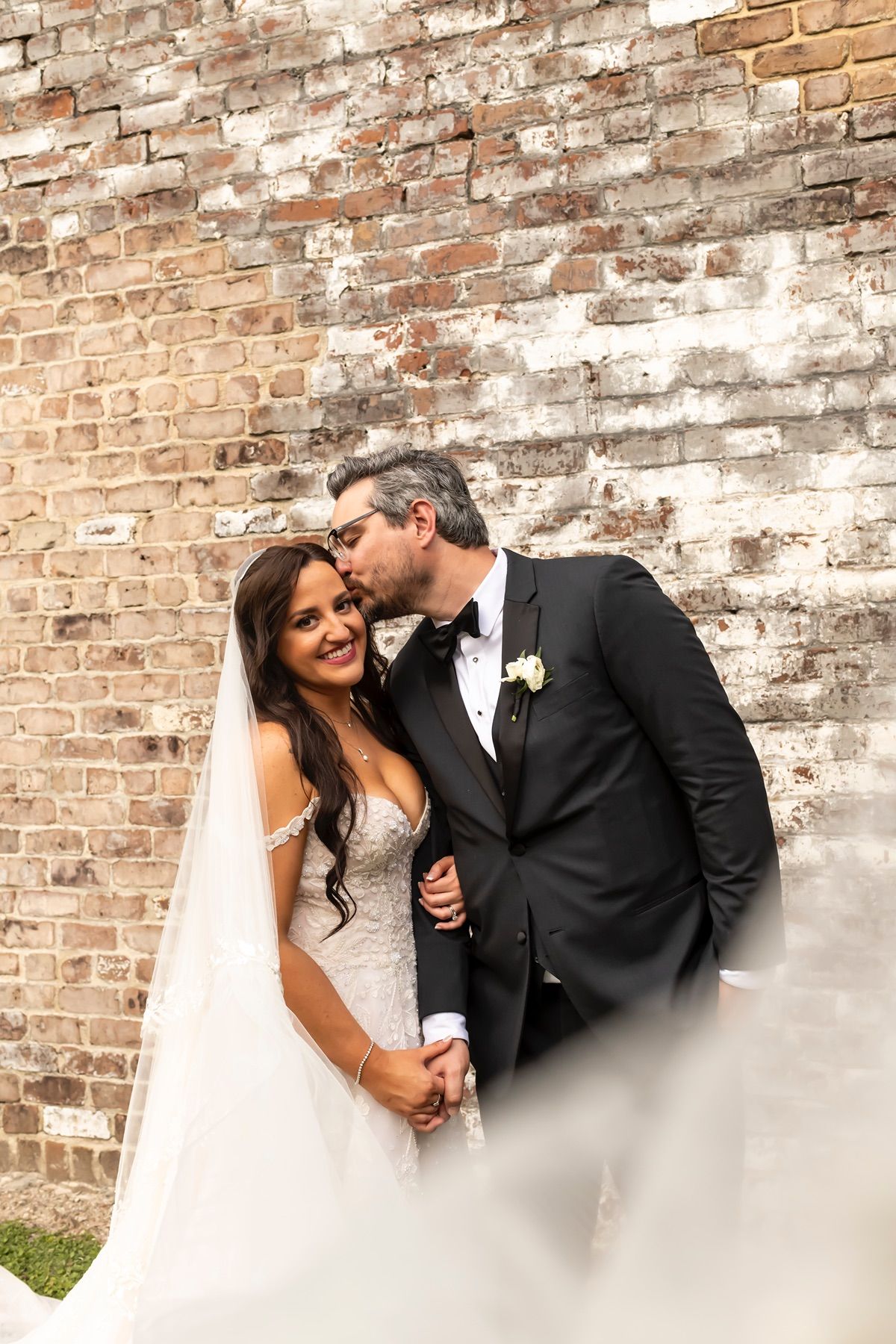 A romantic wedding couple poses together against a weathered brick wall outdoors.