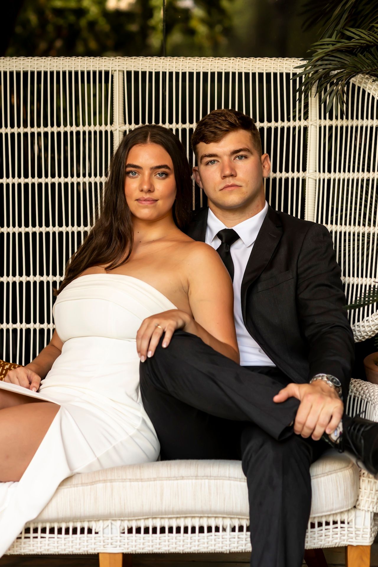 A bride and groom pose together on a white bench in formal wedding attire for romantic outdoor portraits.