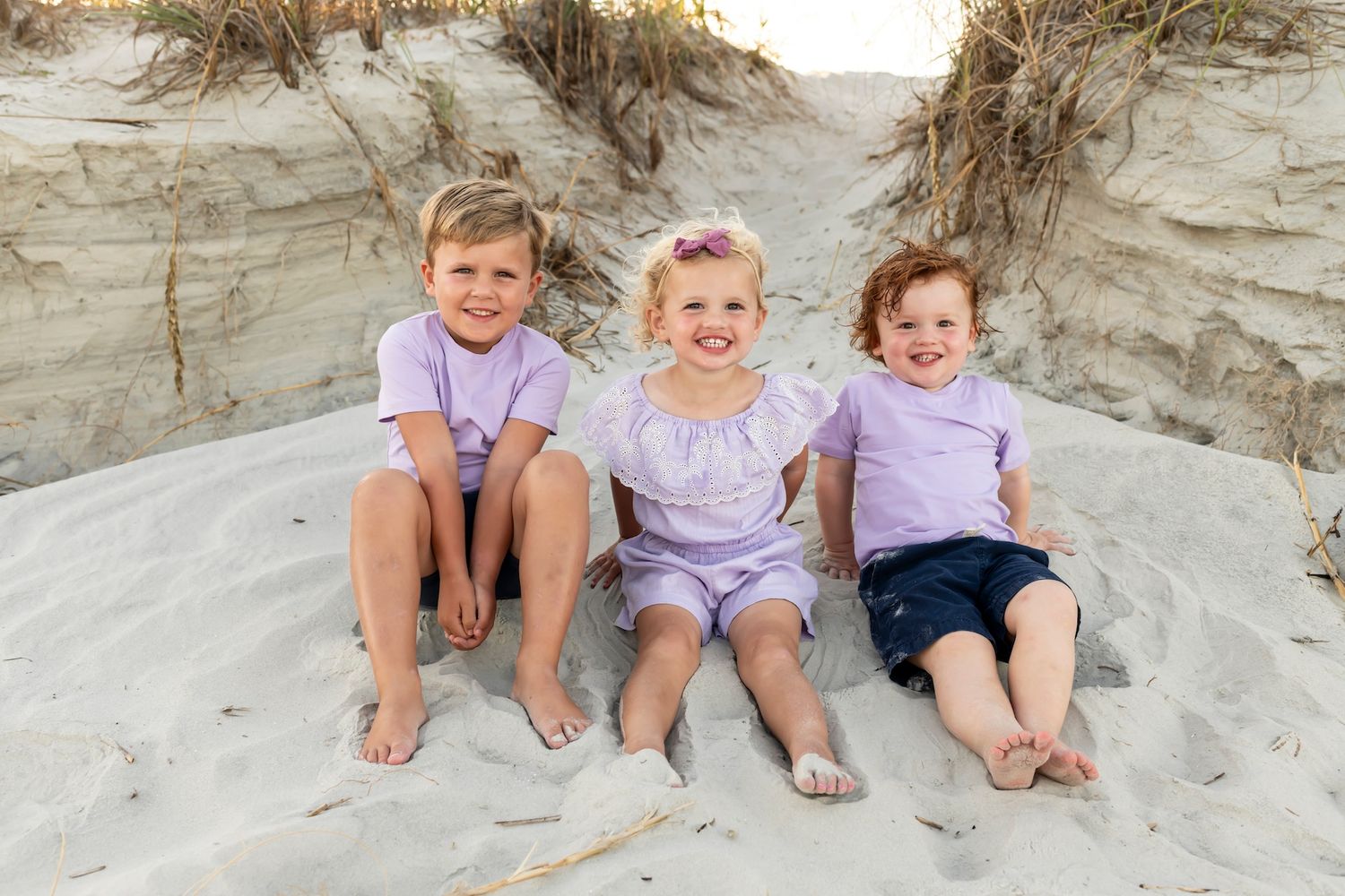 Three children in light purple outfits sit together on a sandy beach dune with grasses behind them.