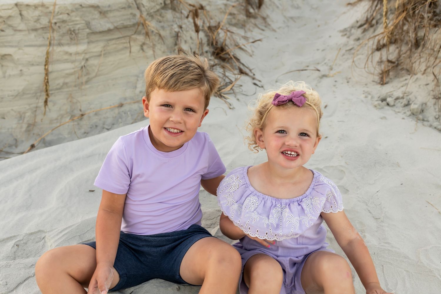 Two children sit smiling together on a sunny beach with sand dunes in the background.
