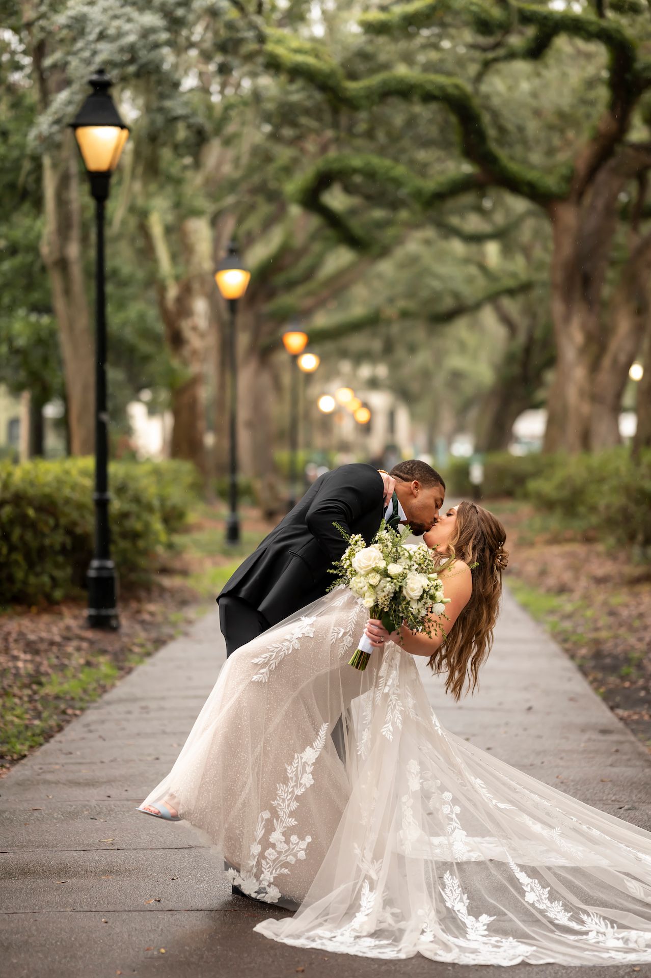 A romantic scene unfolds on a tree-lined pathway with a couple in formal wedding attire sharing intimate moments.