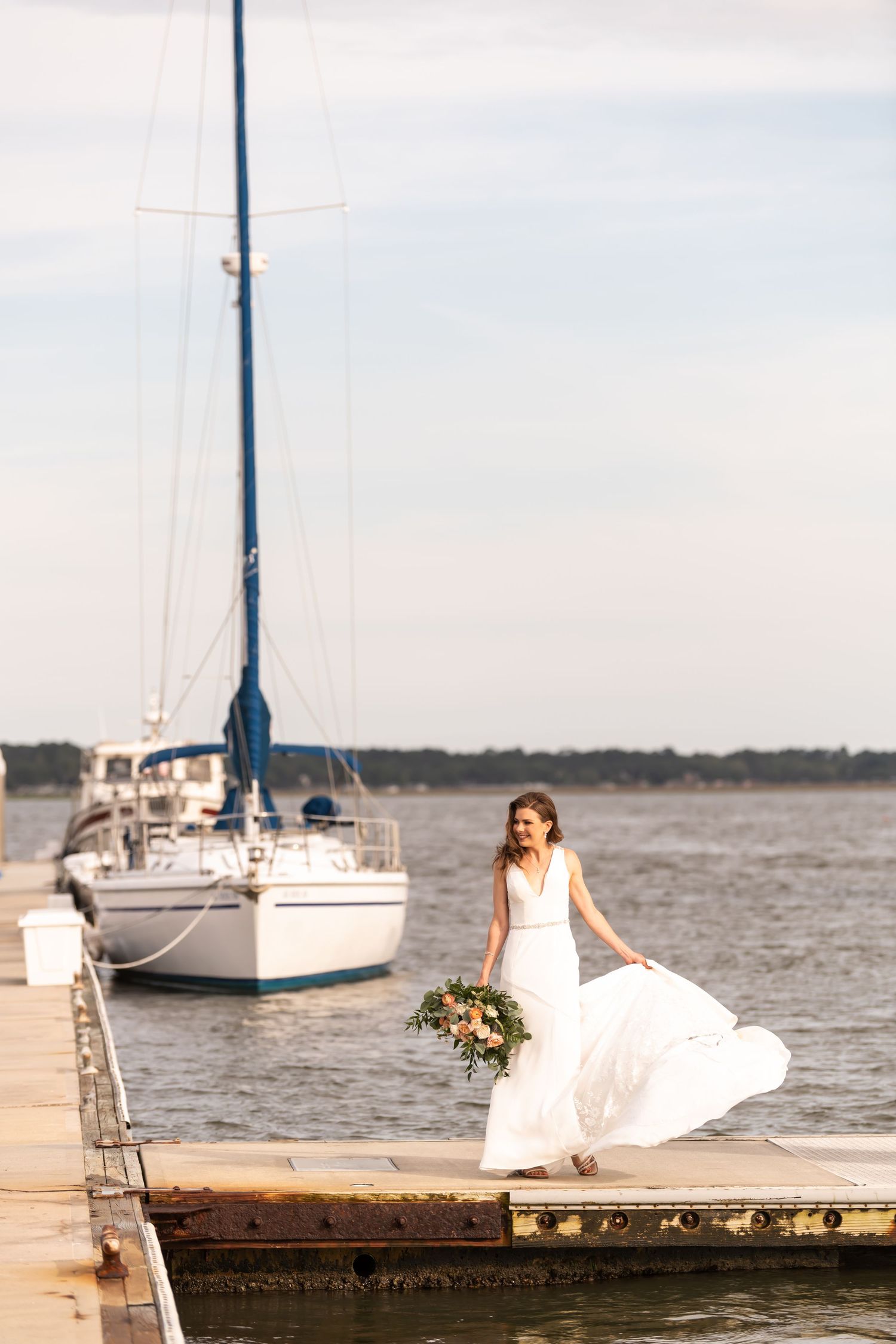 A bride in a flowing white dress poses on a dock with a sailboat in the background at Savannah Yacht Club.