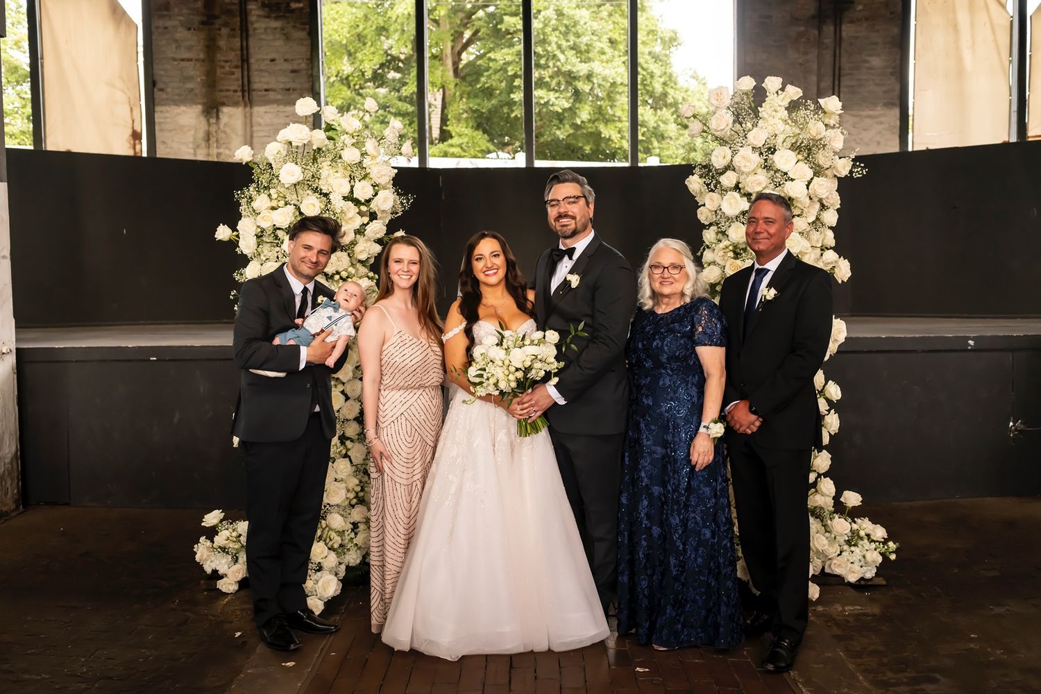 Wedding party poses together for formal photos in an elegant indoor venue with white floral arrangements.