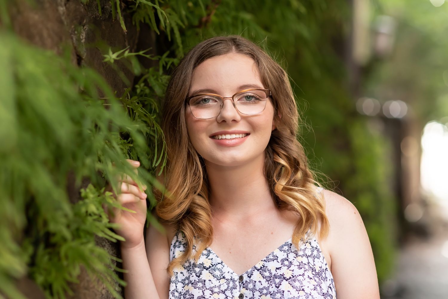 Smiling young woman in floral print dress and glasses stands outdoors among green foliage on sunny day.