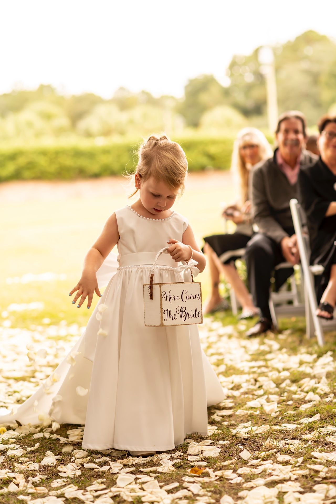 A small child in a white dress scatters flower petals down an aisle during an outdoor golden hour wedding ceremony.