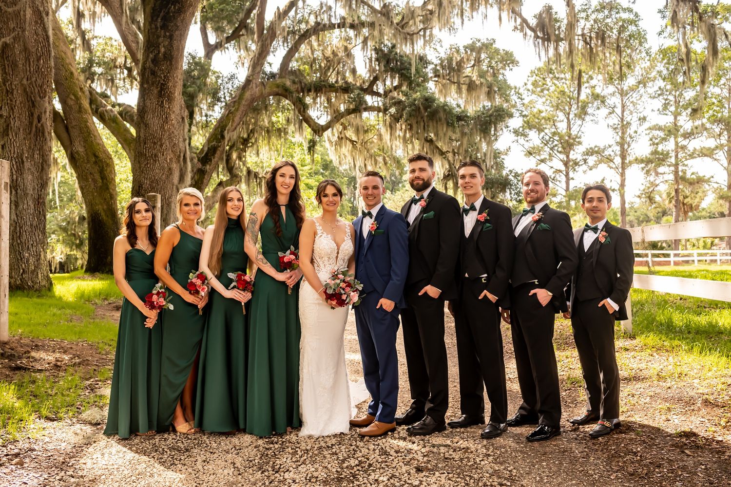 Wedding party poses outdoors under moss-draped oak trees with bridesmaids in green dresses and groomsmen in black suits.
