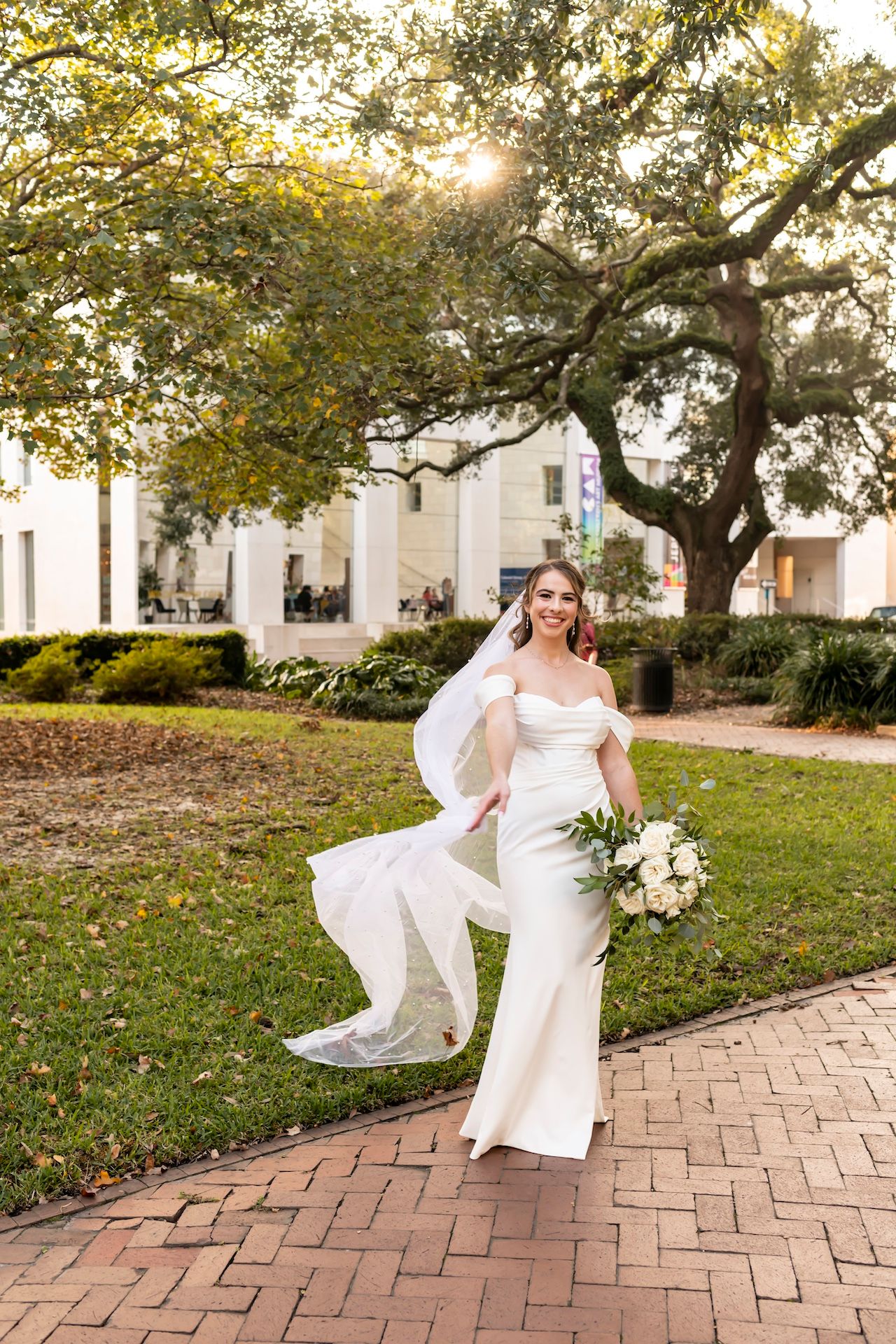 A bride in a fitted white gown and flowing veil walks along a brick path under a majestic oak tree during golden hour.