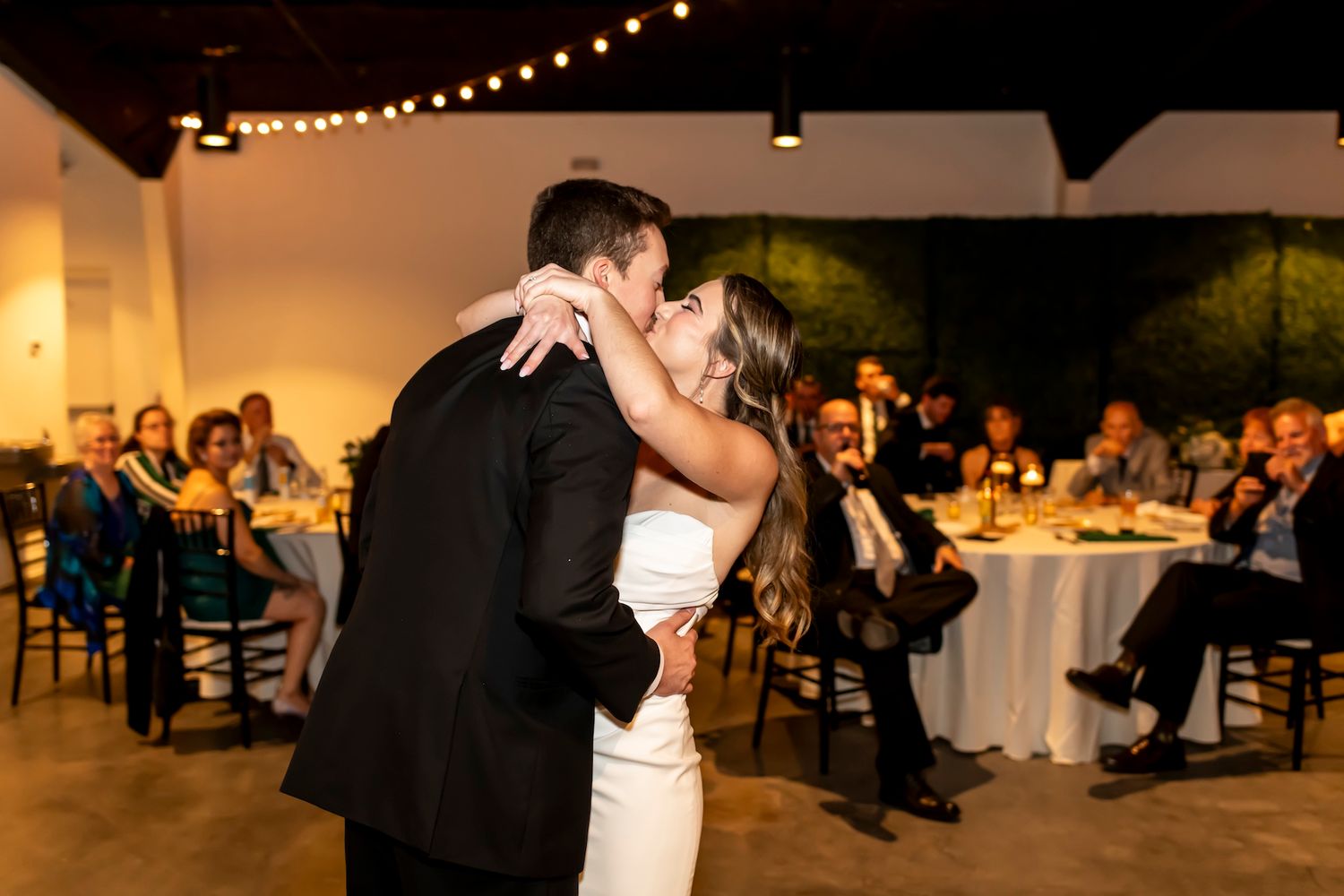 A couple shares their first dance at a wedding reception with string lights and guests seated at tables in the background.
