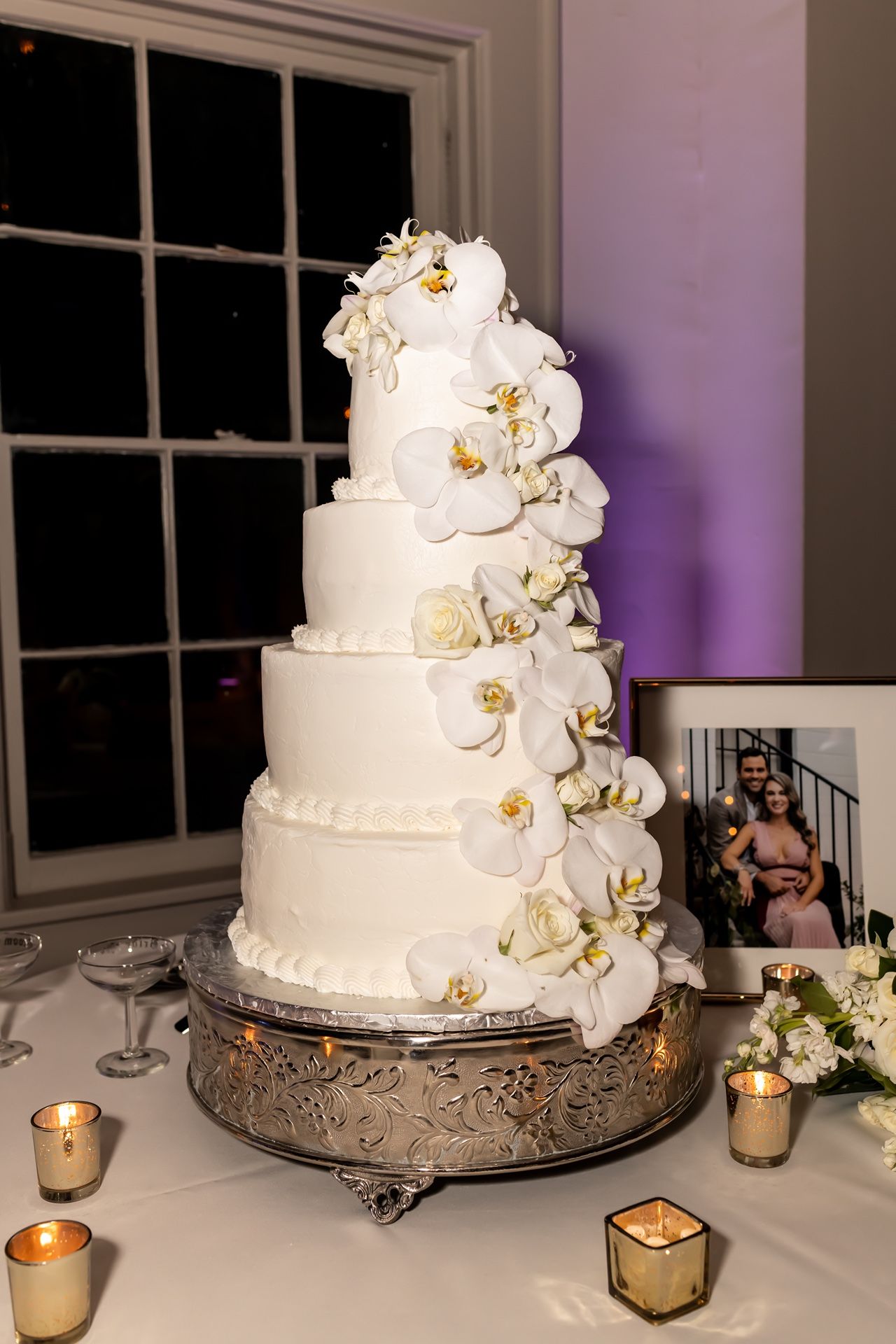 White tiered wedding cake decorated with delicate orchids and displayed on an ornate silver stand with glowing candles nearby.