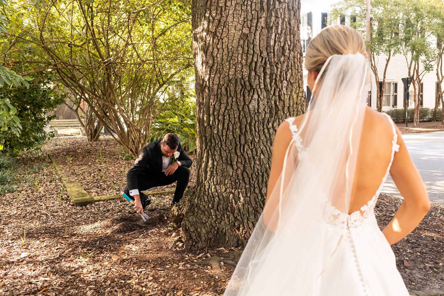 A photographer crouches behind a tree to capture a dramatic wedding photo in a sunlit garden setting.